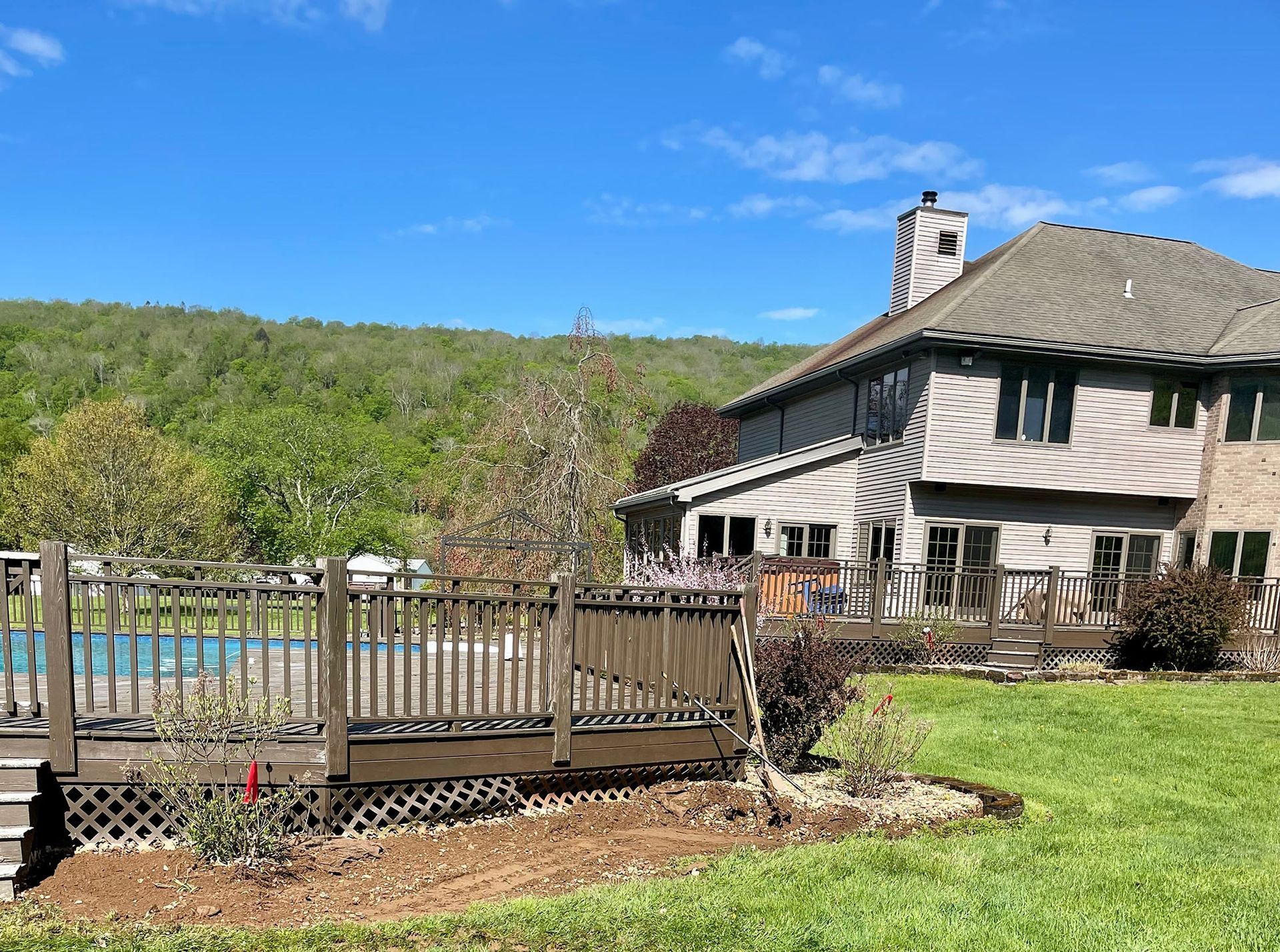 Back of house with deck and pool overlooking a green hillside under a blue sky.
