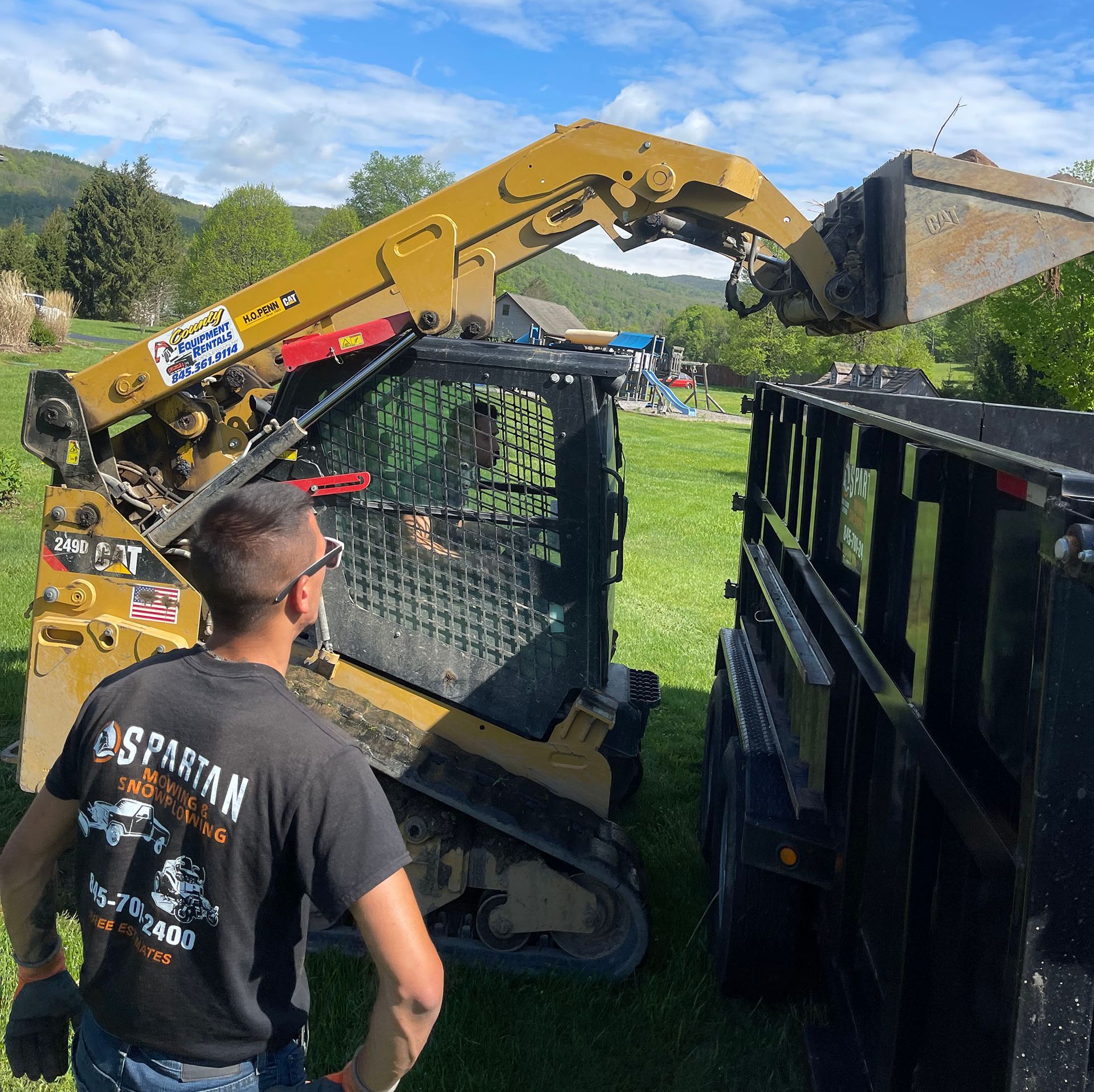 Man operating skid steer, loading debris into a trailer in a grassy field.