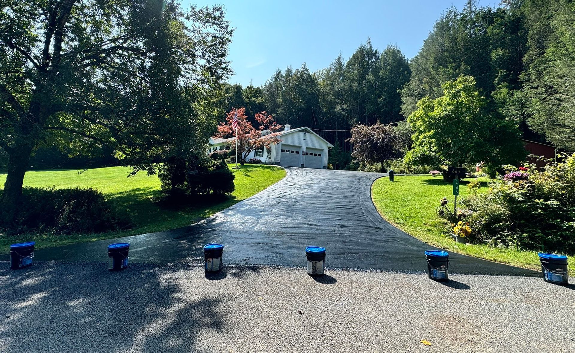 Black asphalt driveway leading to a white house with a garage, flanked by green lawns and trees under a blue sky.