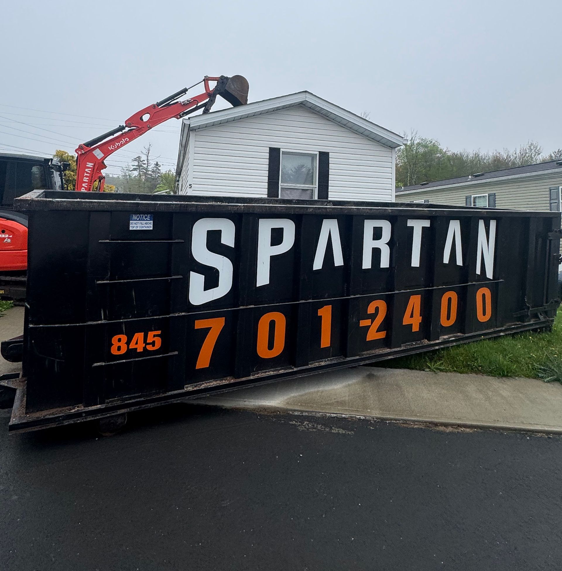 Black Spartan dumpster with white and orange lettering; 845-701-2400. Excavator in background.