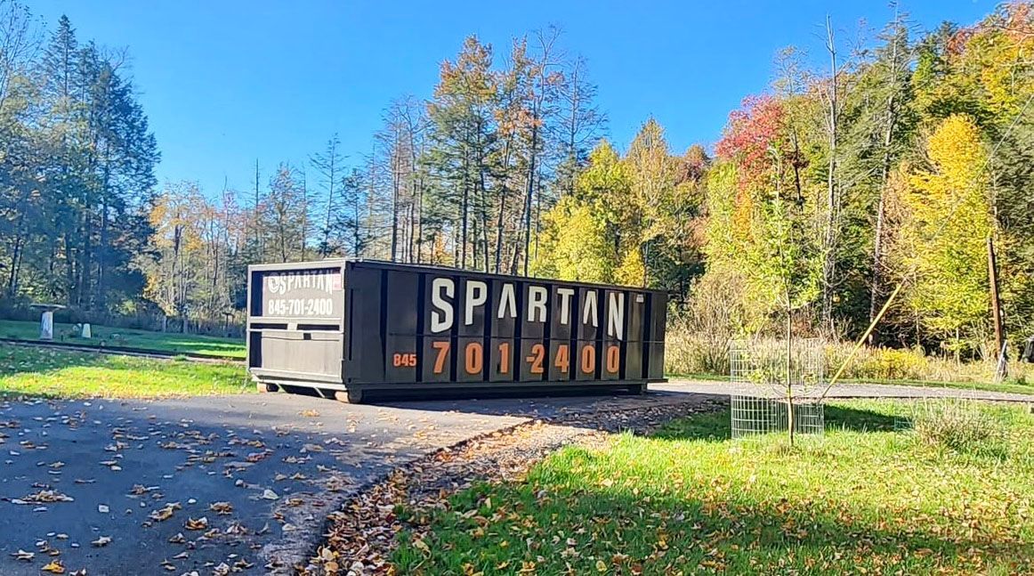 Large Spartan dumpster on a paved driveway, surrounded by fall foliage under a clear blue sky.