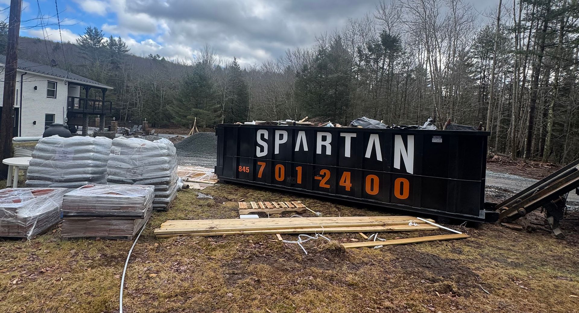 Black Spartan dumpster with phone number, in front of a house, construction materials nearby.