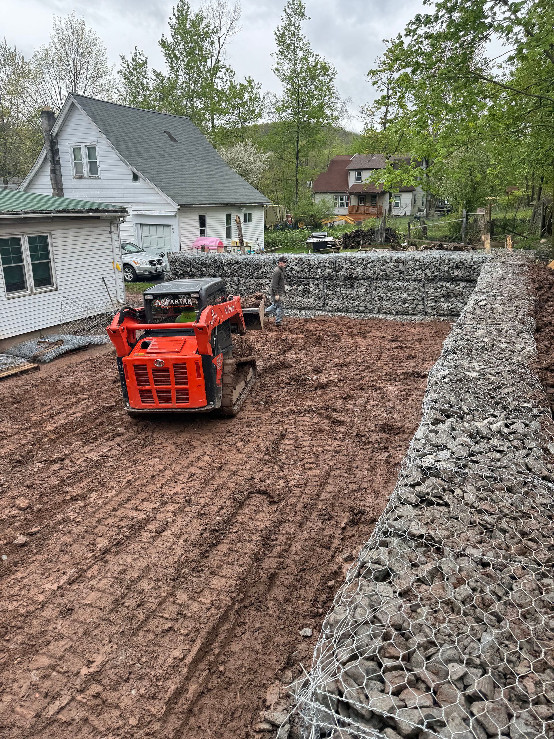 An orange skid steer compacting red mud in a construction site with gabion walls and houses in the background.