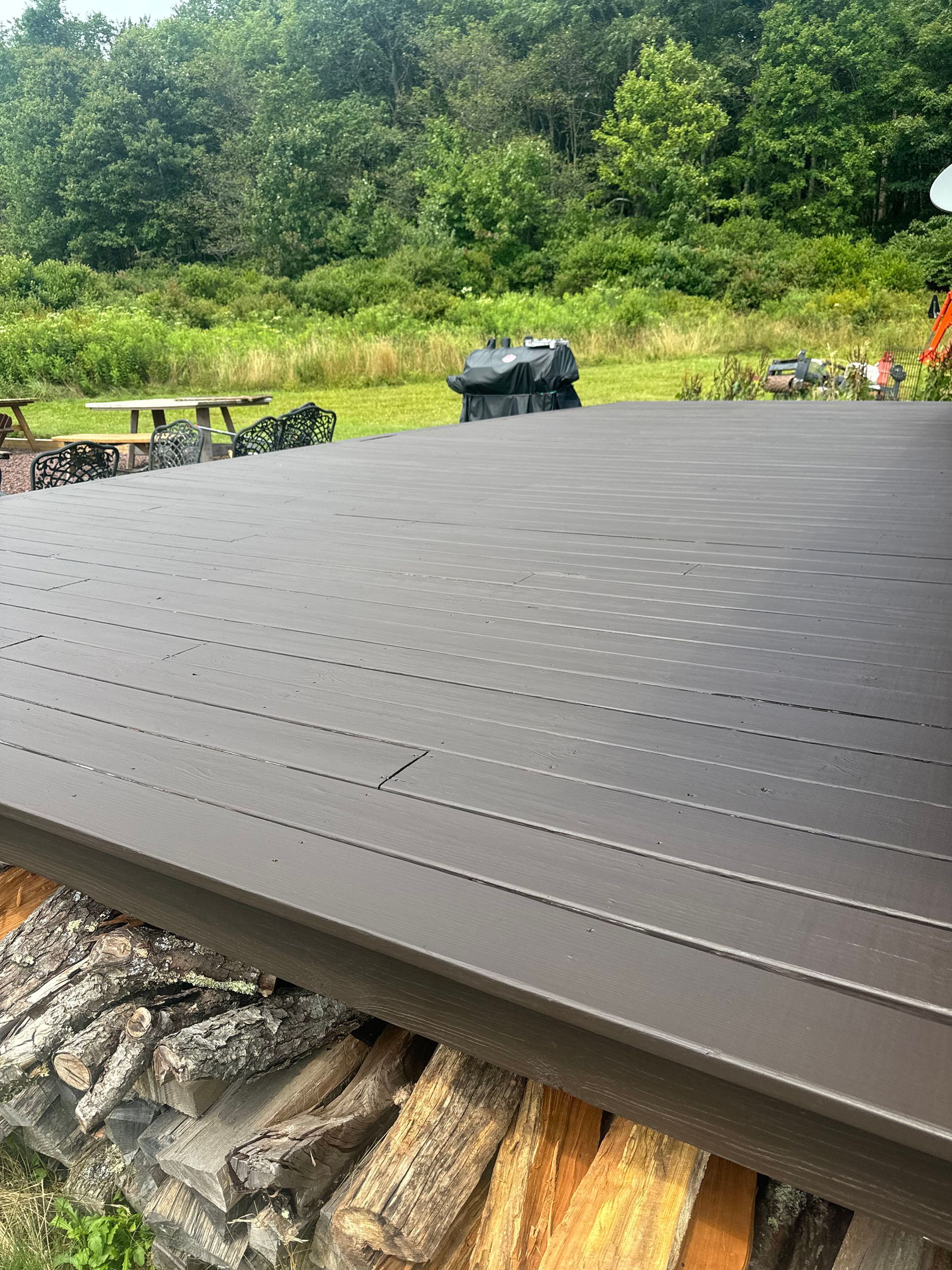 Brown metal patio cover over a firewood stack, with a grill and trees in the background.