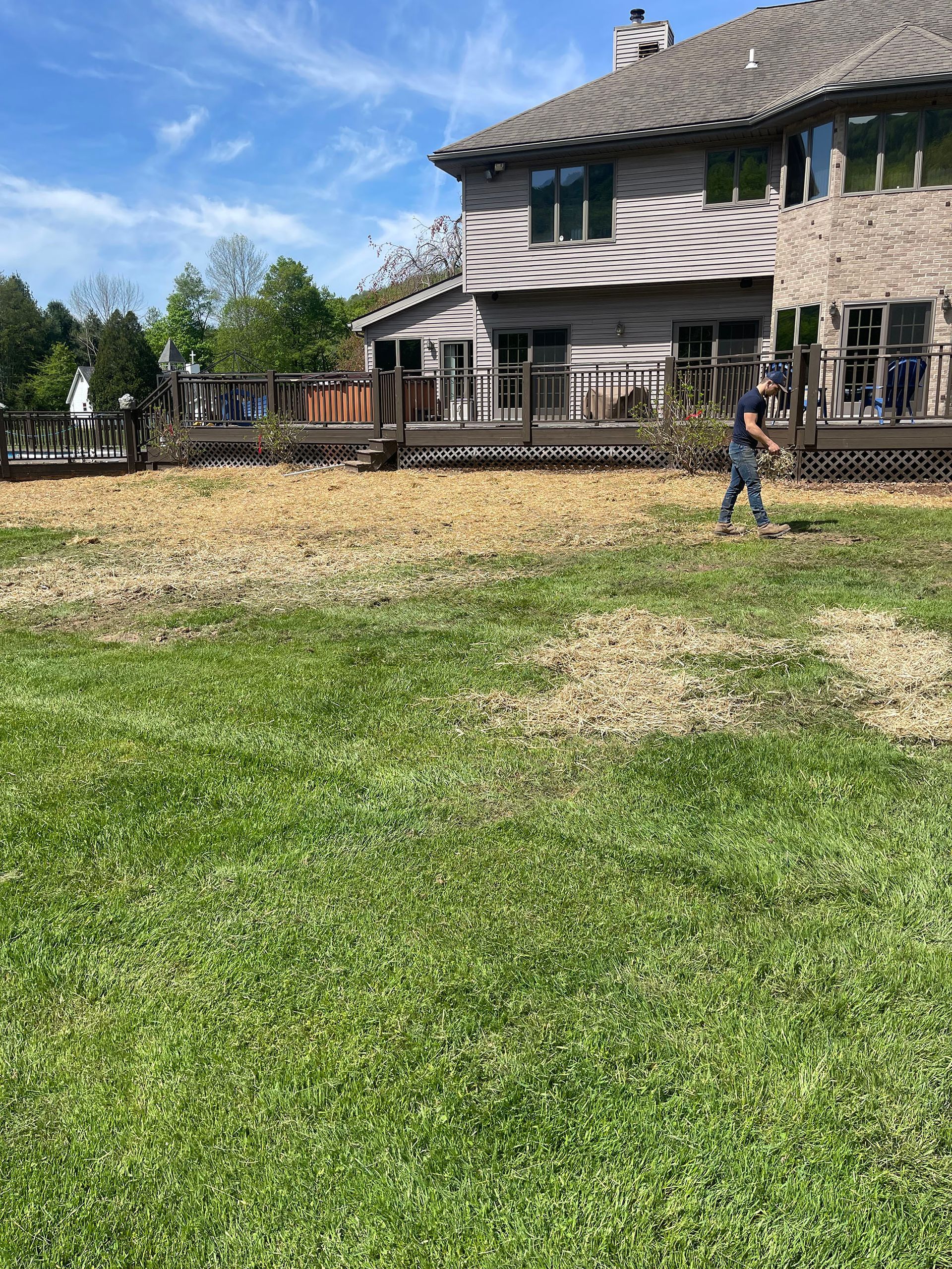 Man working on a patchy lawn in front of a house with a large deck; sunny day.