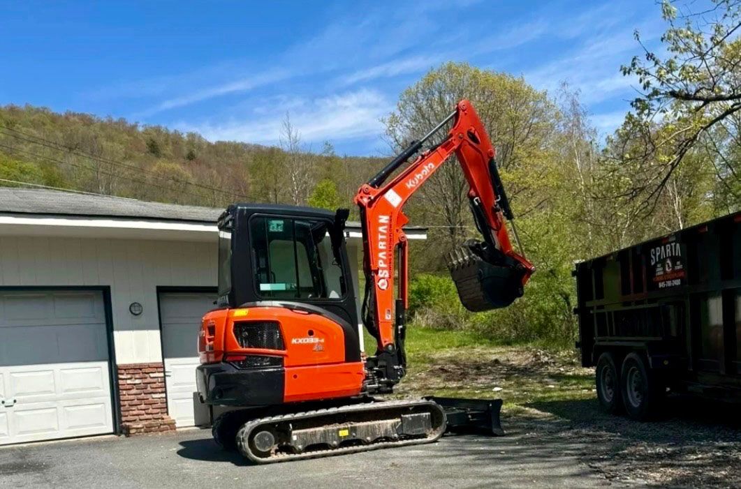 Orange and black excavator next to a garage, near a trailer. Blue sky, trees in the background.