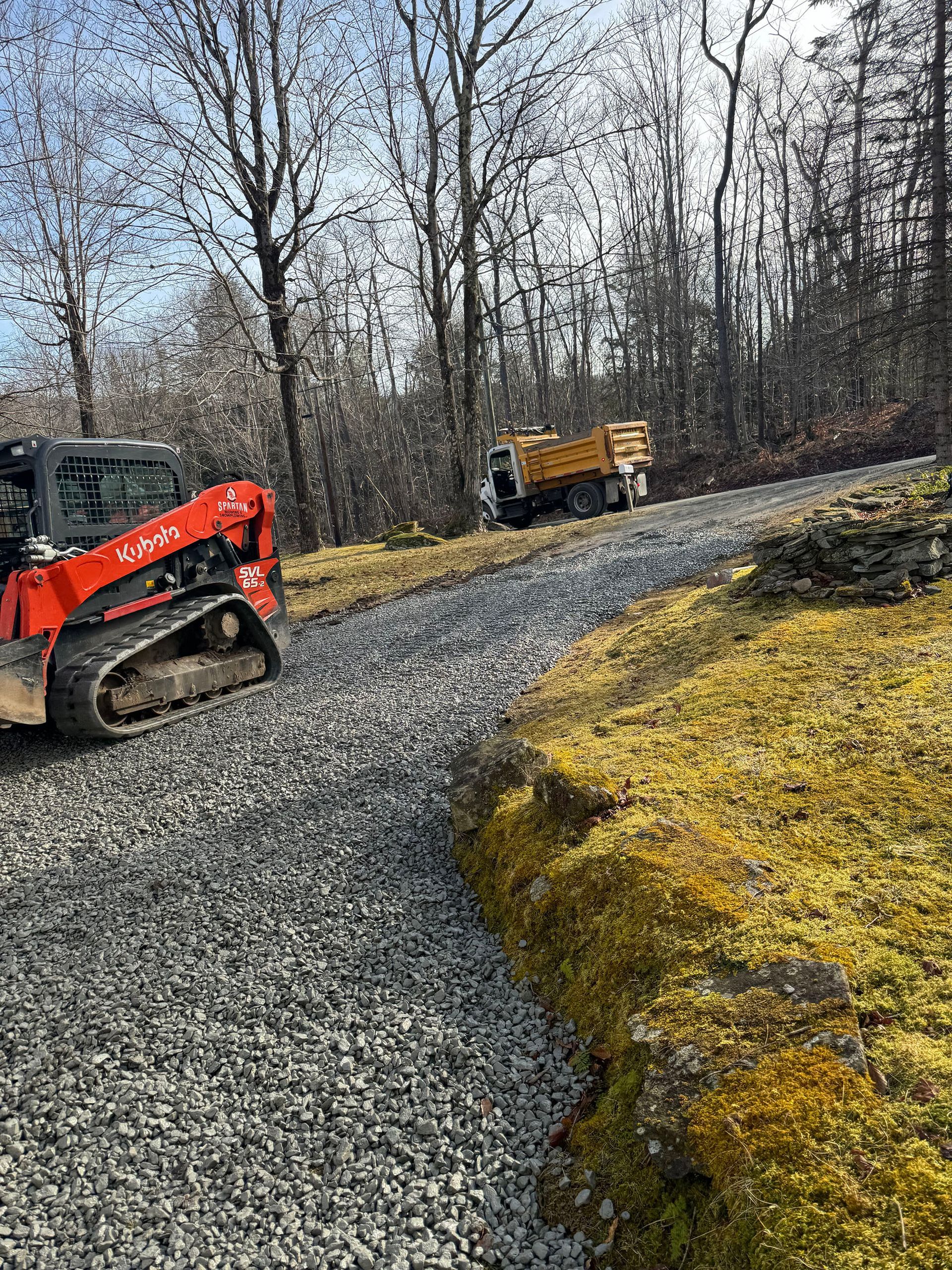 A gravel driveway under construction, with an orange skid steer and a dump truck.