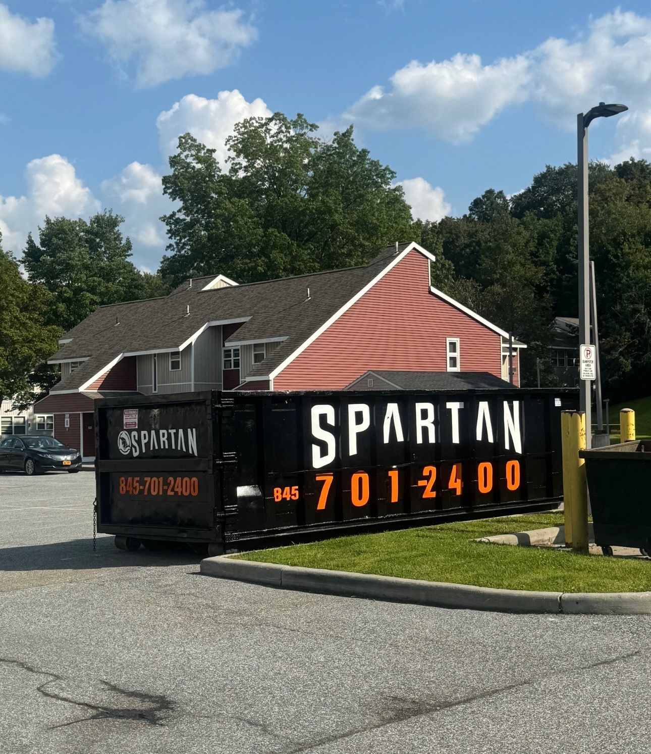 Black Spartan dumpster in a parking area with a red-sided building, blue sky, and trees in the background.