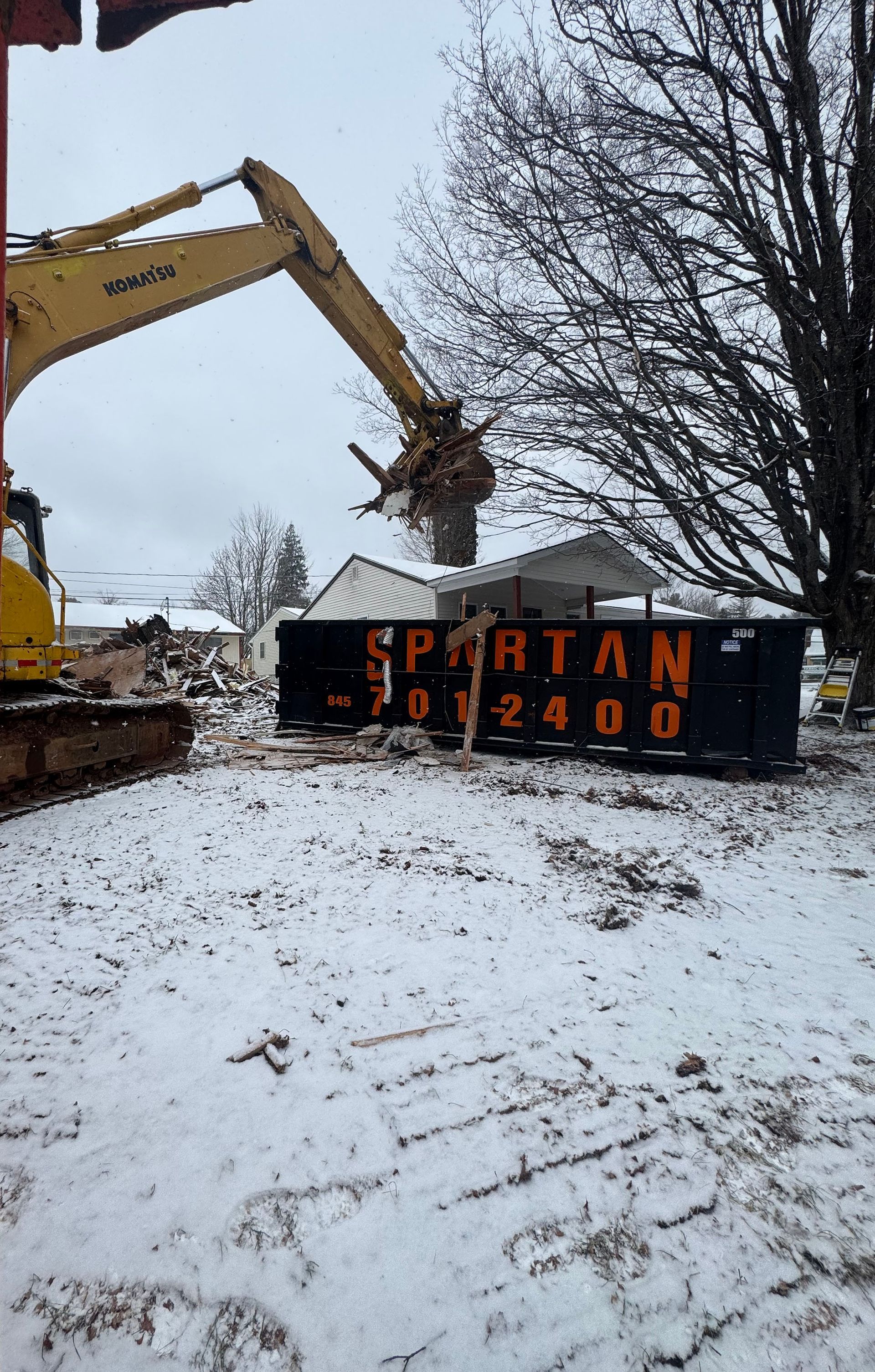An excavator dumps debris into a Spartan dumpster in a snowy residential area.