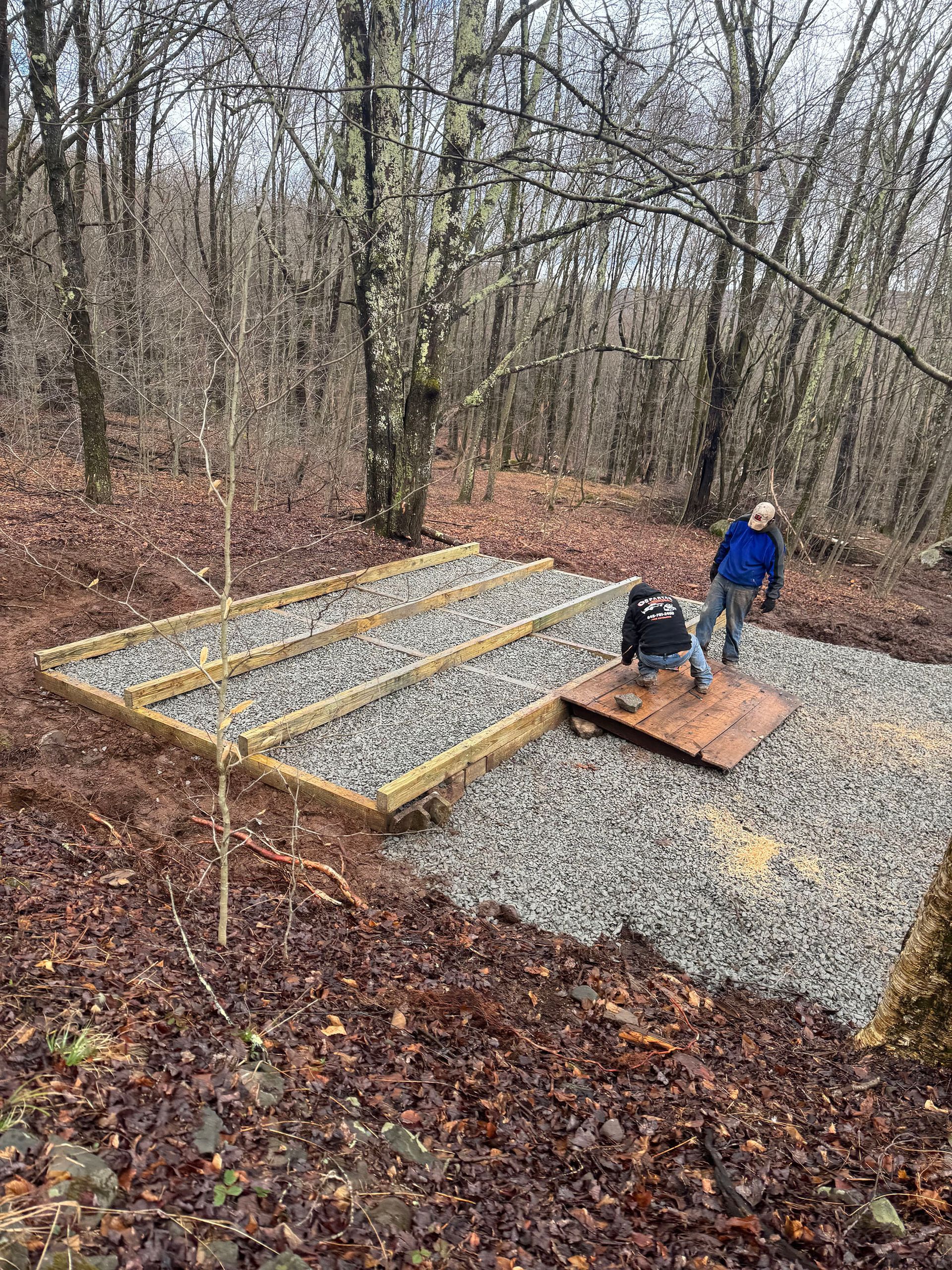 Gravel-covered construction site in a wooded area. Man working, wood frame, and a dark box. Brown leaves and trees.
