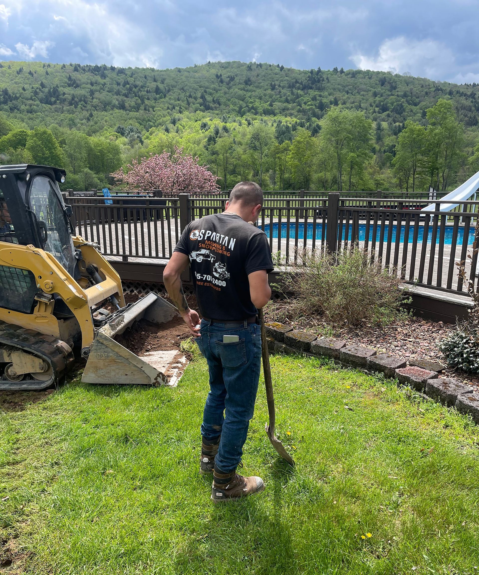 Man using a shovel in a yard next to a pool and skid steer, mountains in the background.