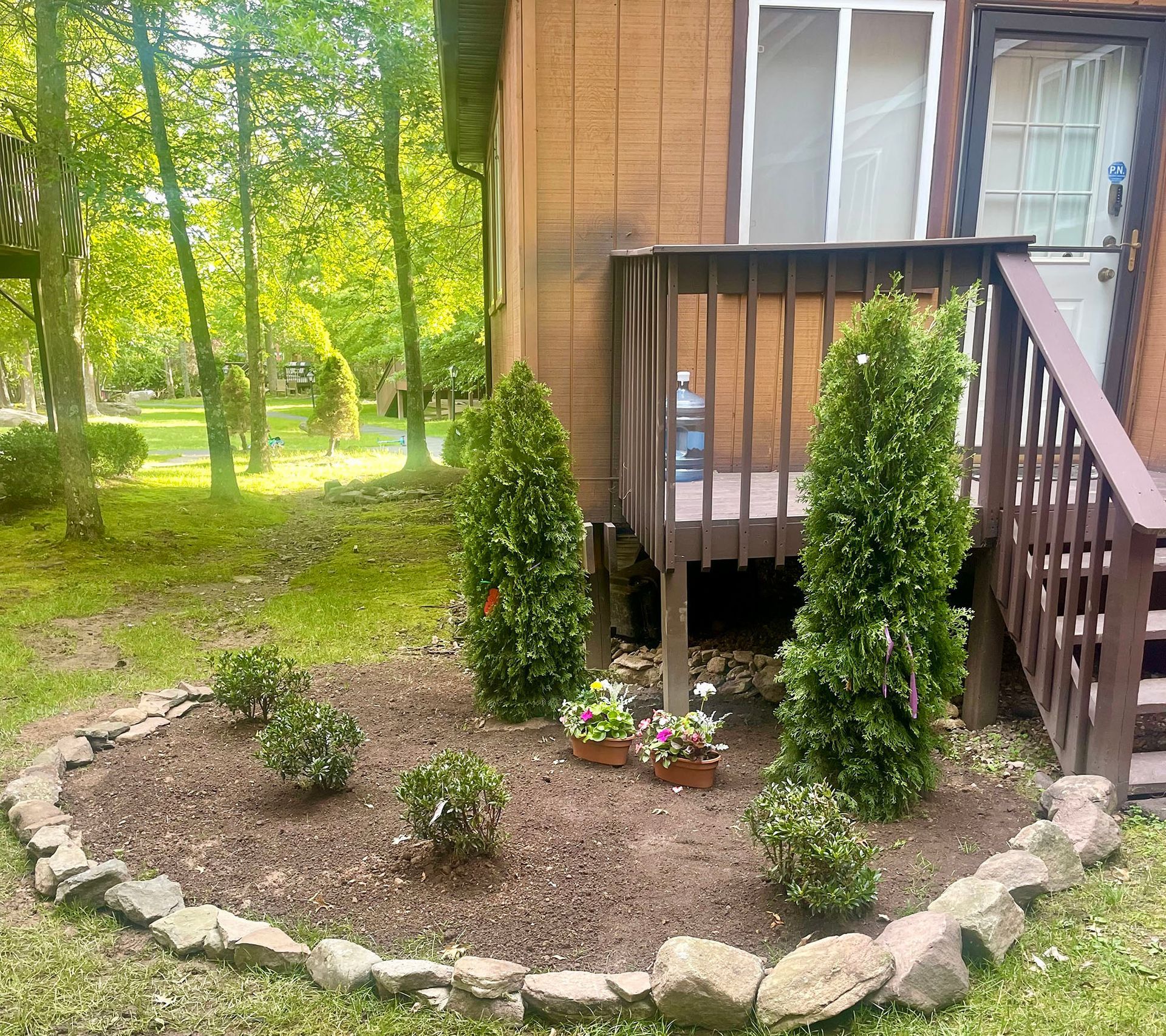 A small garden bed with various plants and rocks in front of a house's wooden deck.