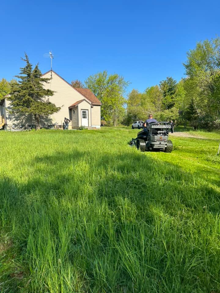 A small, beige house with a long, green lawn; a person mows the grass with a riding mower on a sunny day.