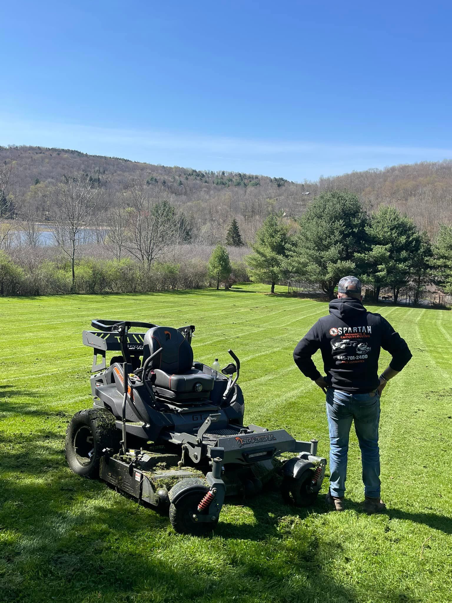 Man standing next to a zero-turn lawnmower on a green lawn with a scenic backdrop of a lake and mountains.