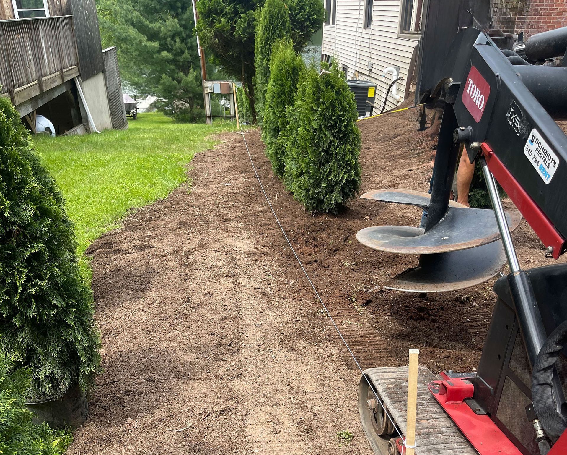 A row of green shrubs in mulch. A red Toro machine with an auger drill. Green grass and a building in the background.