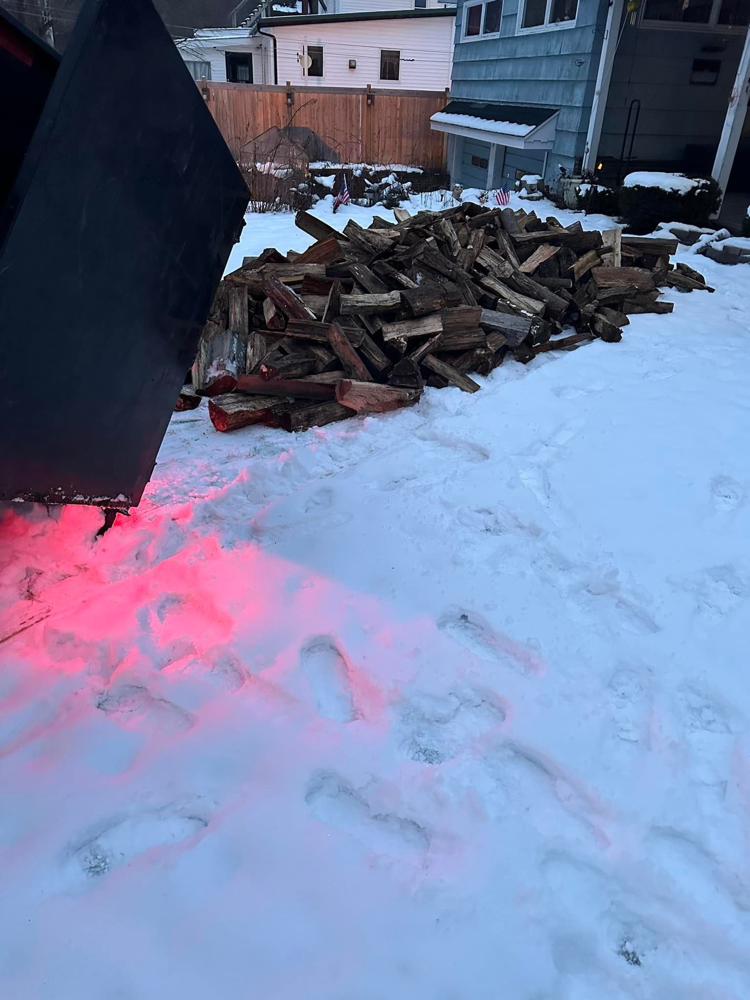 Pile of firewood on snow next to a dark vehicle with red light, houses in the background.