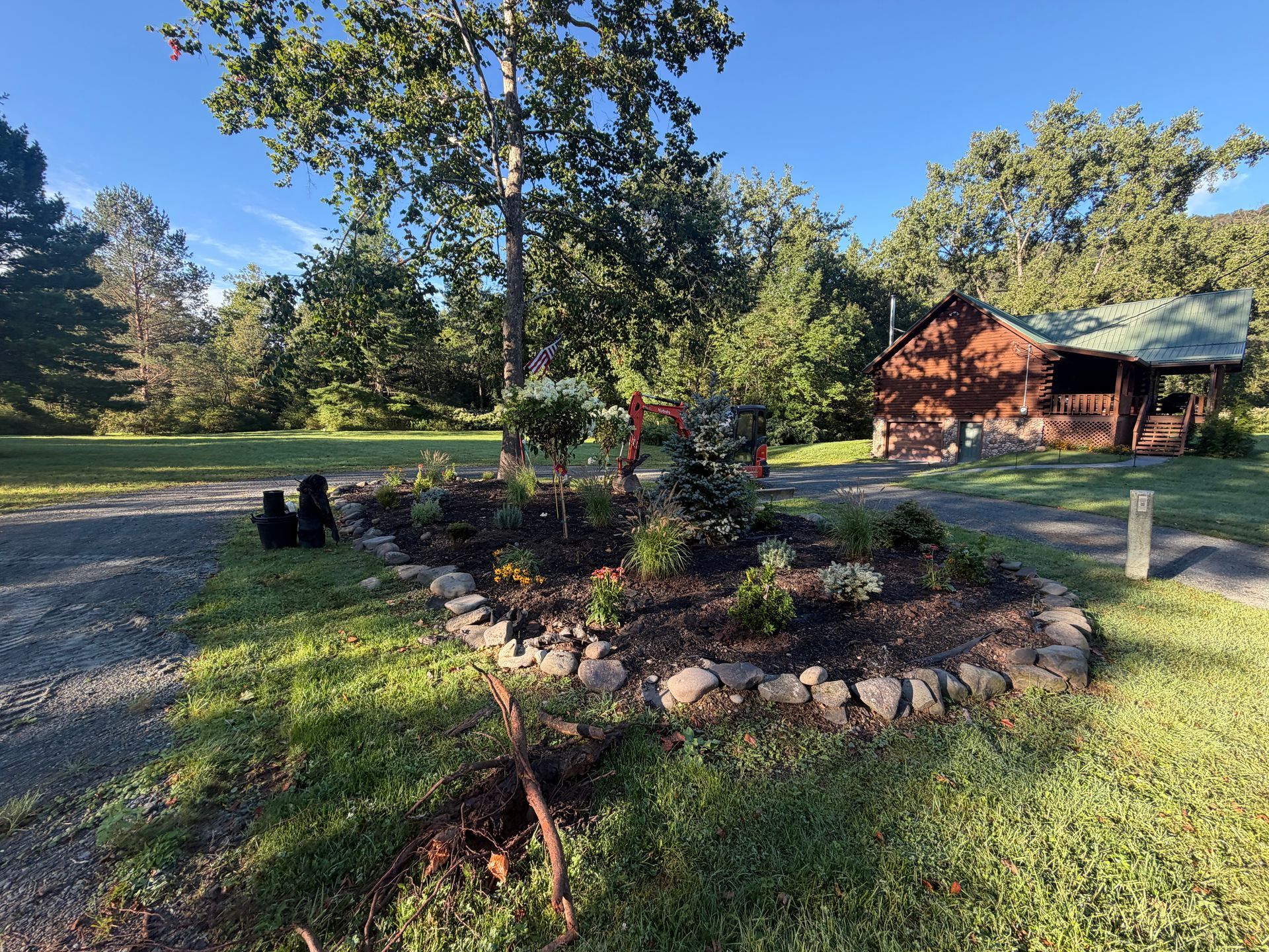Front yard flower bed with red blooms, surrounded by stones beside a gravel drive and a house in the background