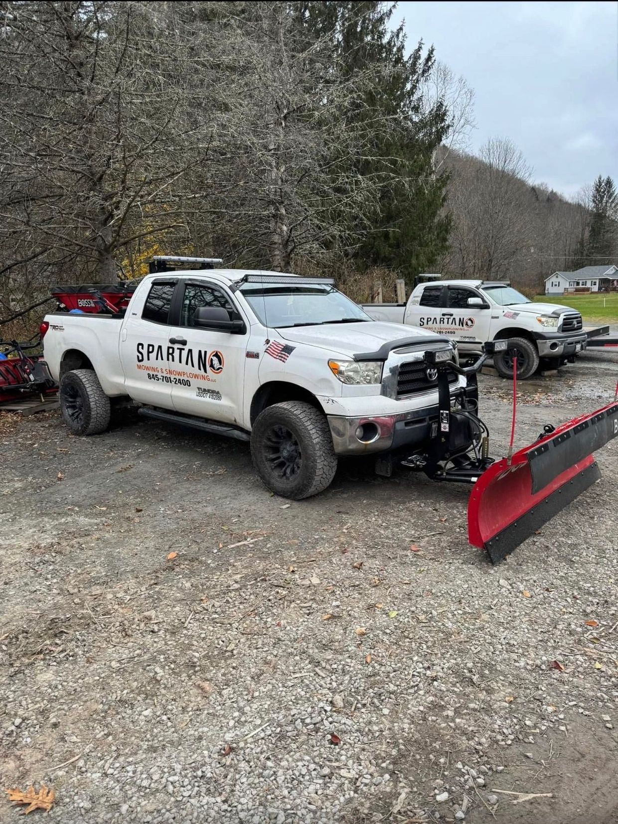White pickup truck with snowplow, parked on gravel, another truck in background. 
