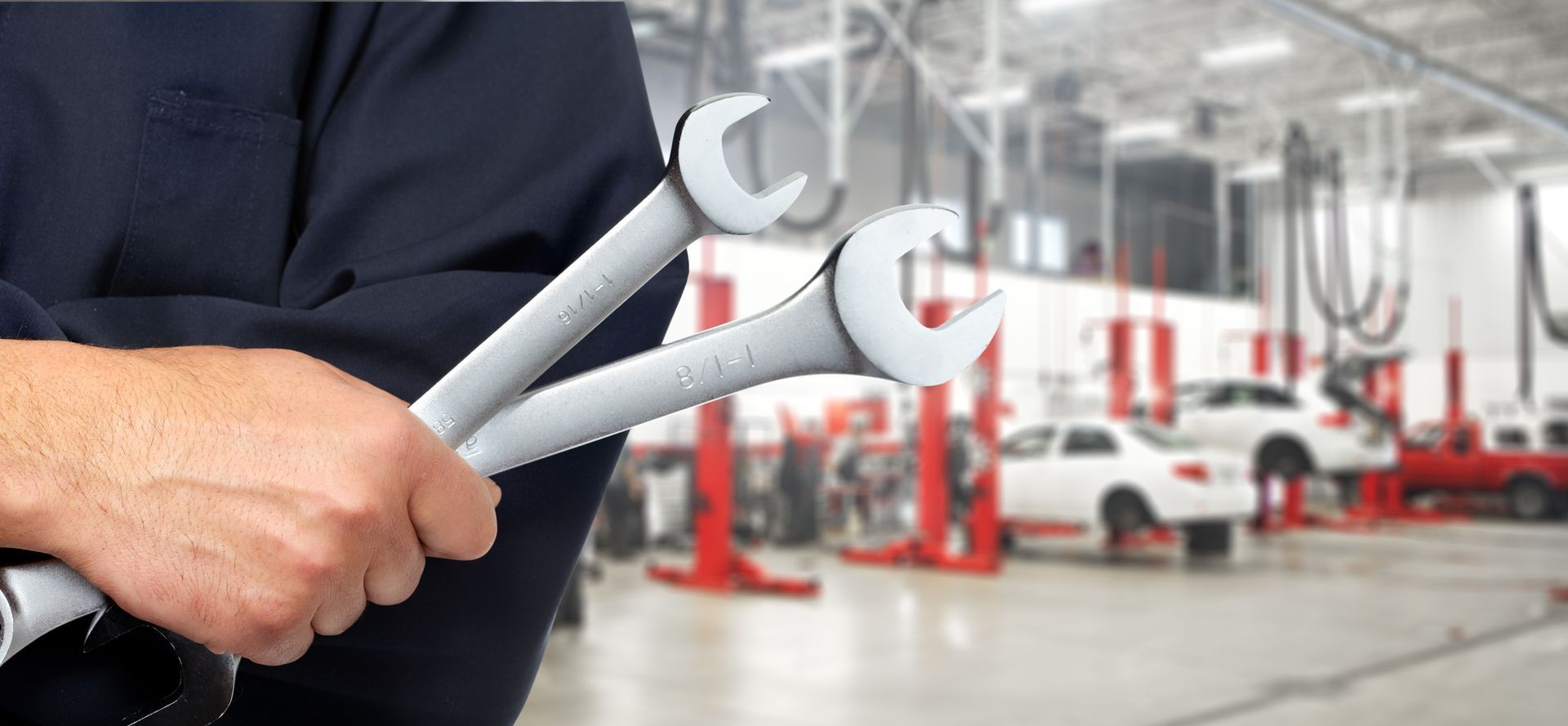Mechanic holding two wrenches in an auto repair shop with cars on lifts in the background.
