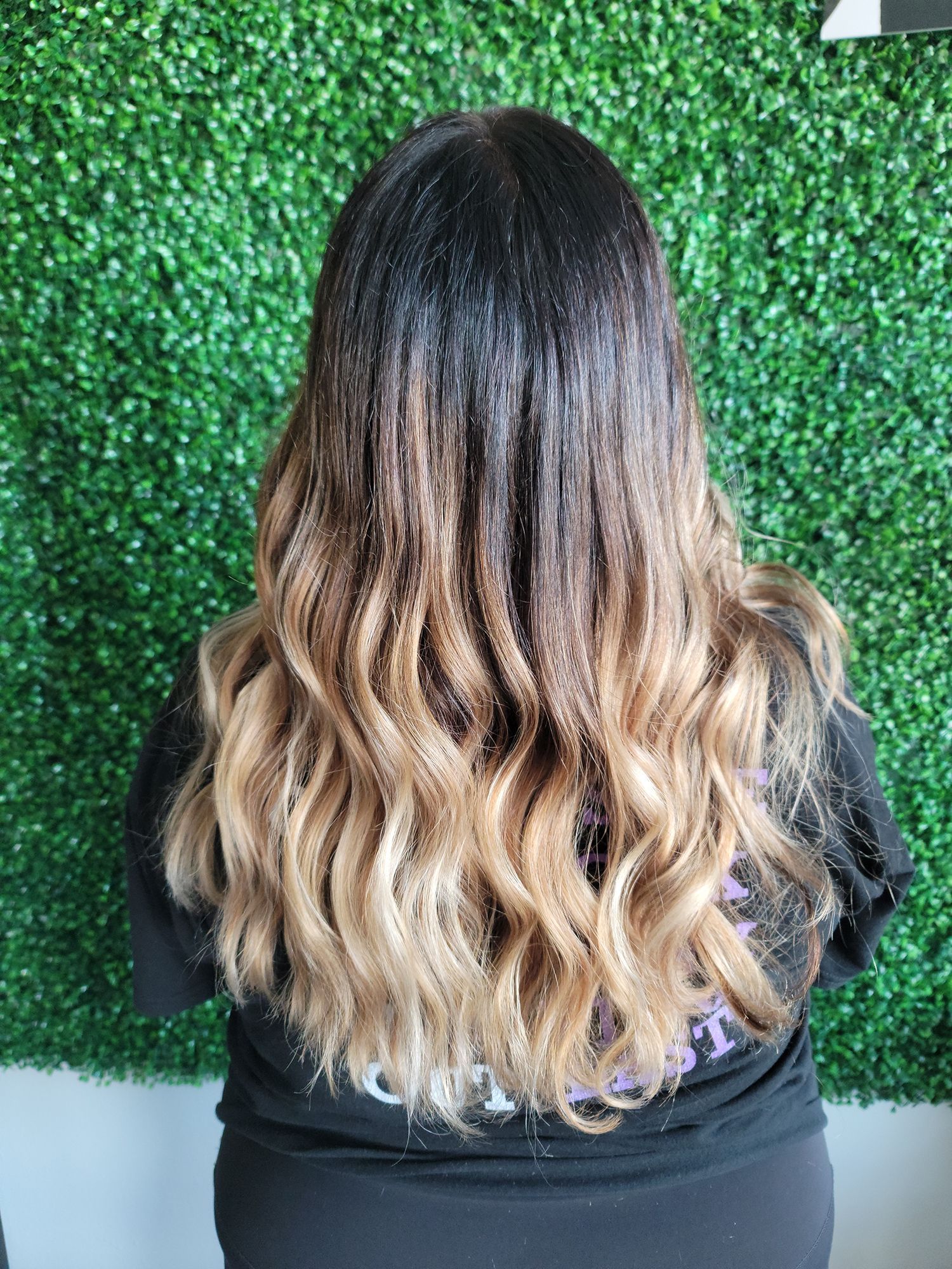A woman with long wavy hair is standing in front of a hedge wall.