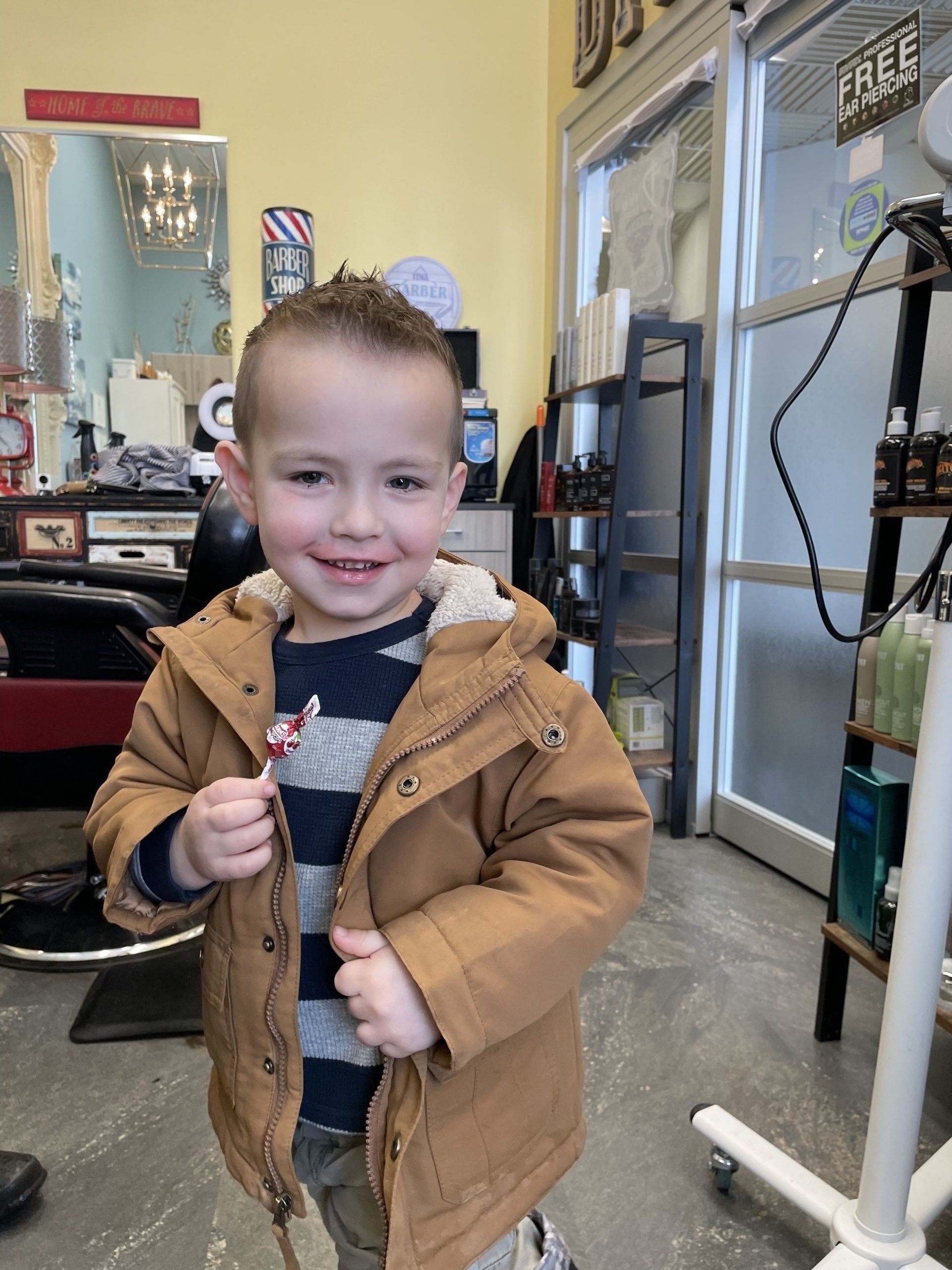 A smiling young boy with a new haircut, wearing a brown coat, standing in a barbershop.