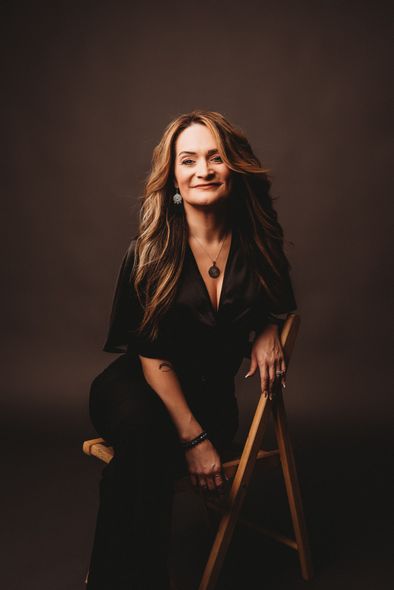 Woman with long brown hair, wearing black dress, sitting on a wooden chair, smiling.