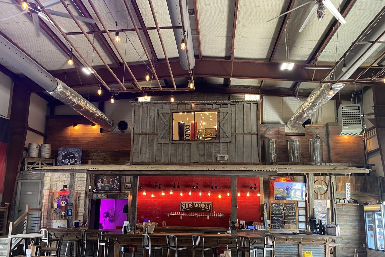 Rustic bar interior with wooden structure, overhead lights, and bar stools. Red wall behind bar.