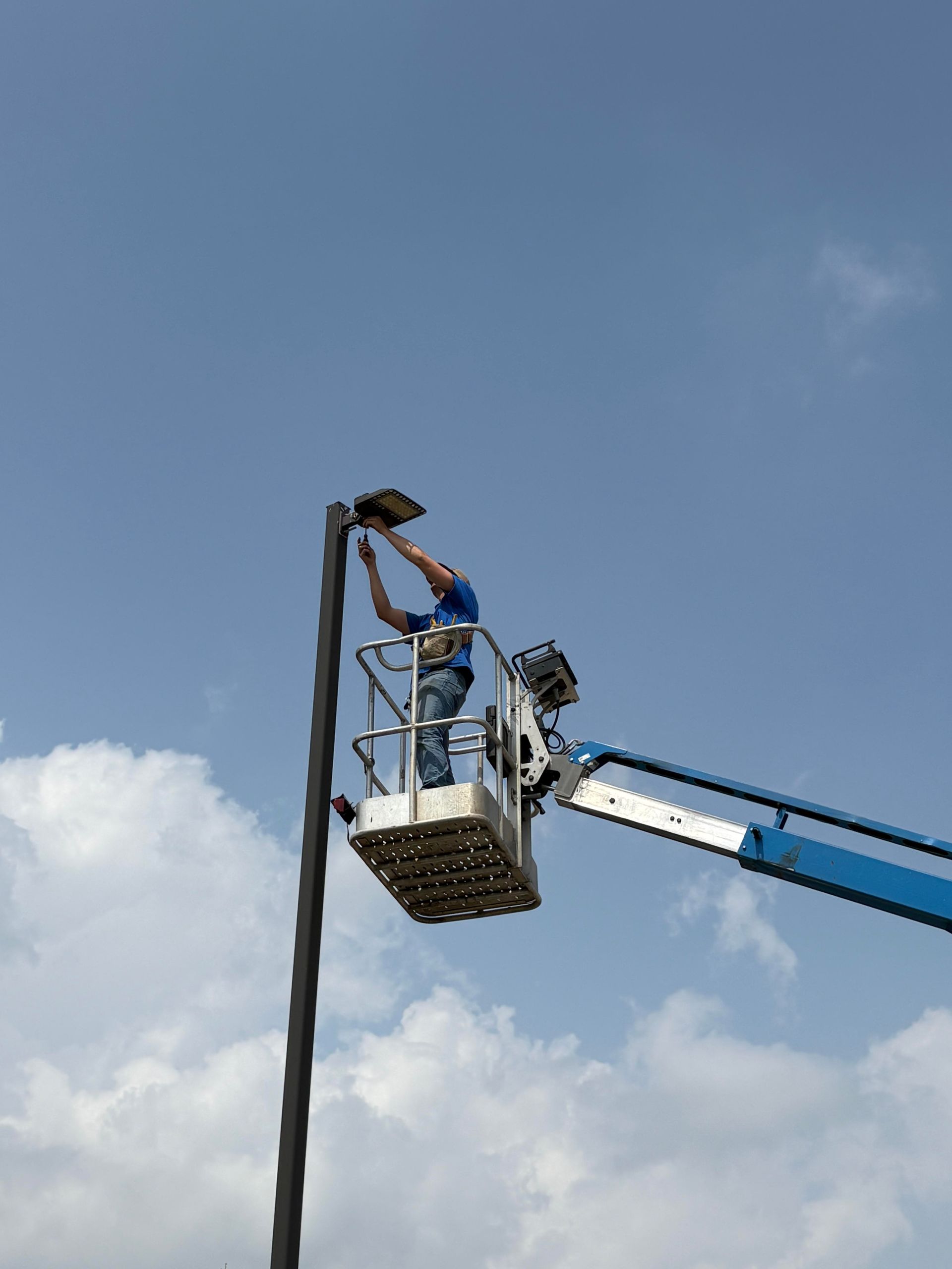 Man in lift installing a light fixture on a pole against a partly cloudy sky.