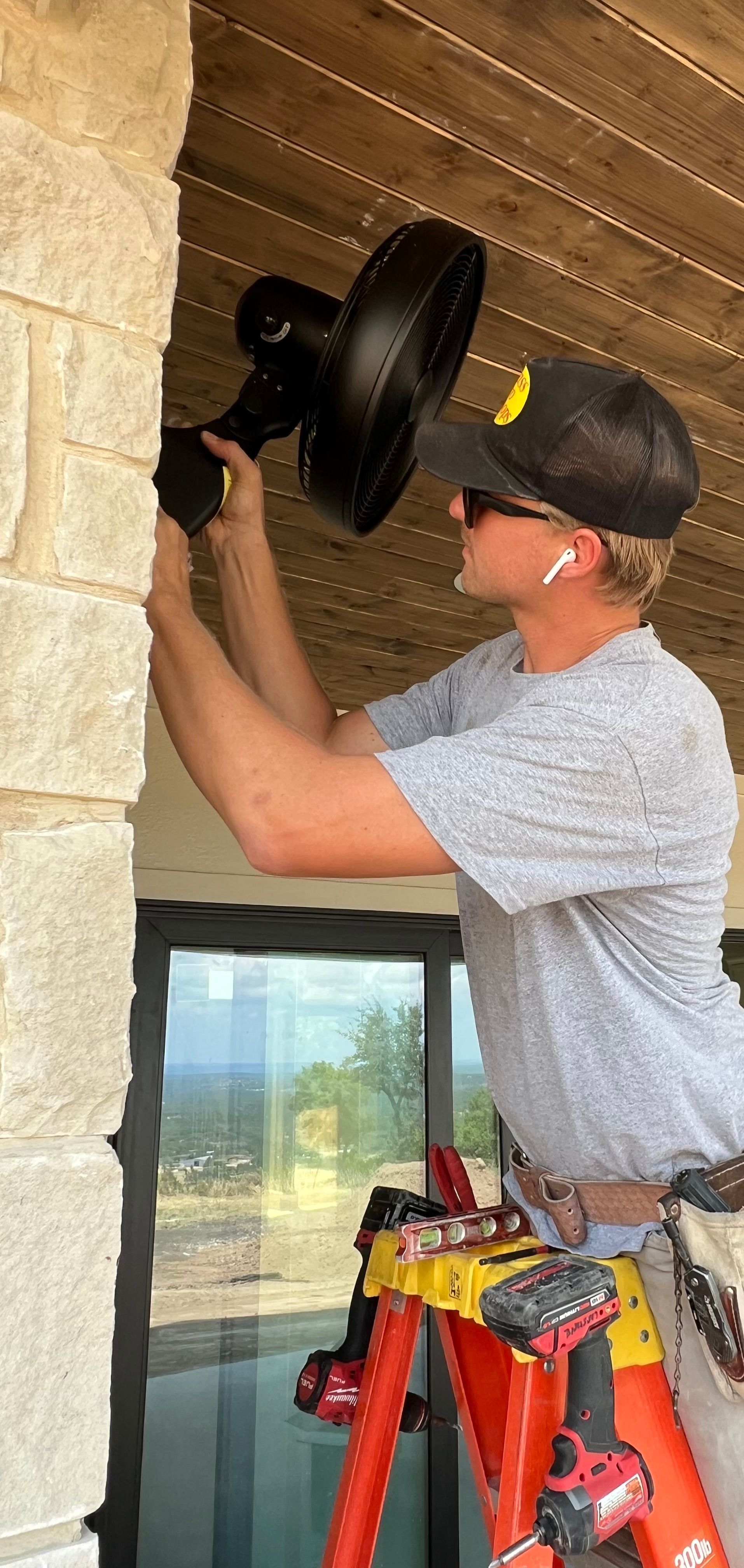 Man installing a black fan on a stone wall, standing on a red ladder.
