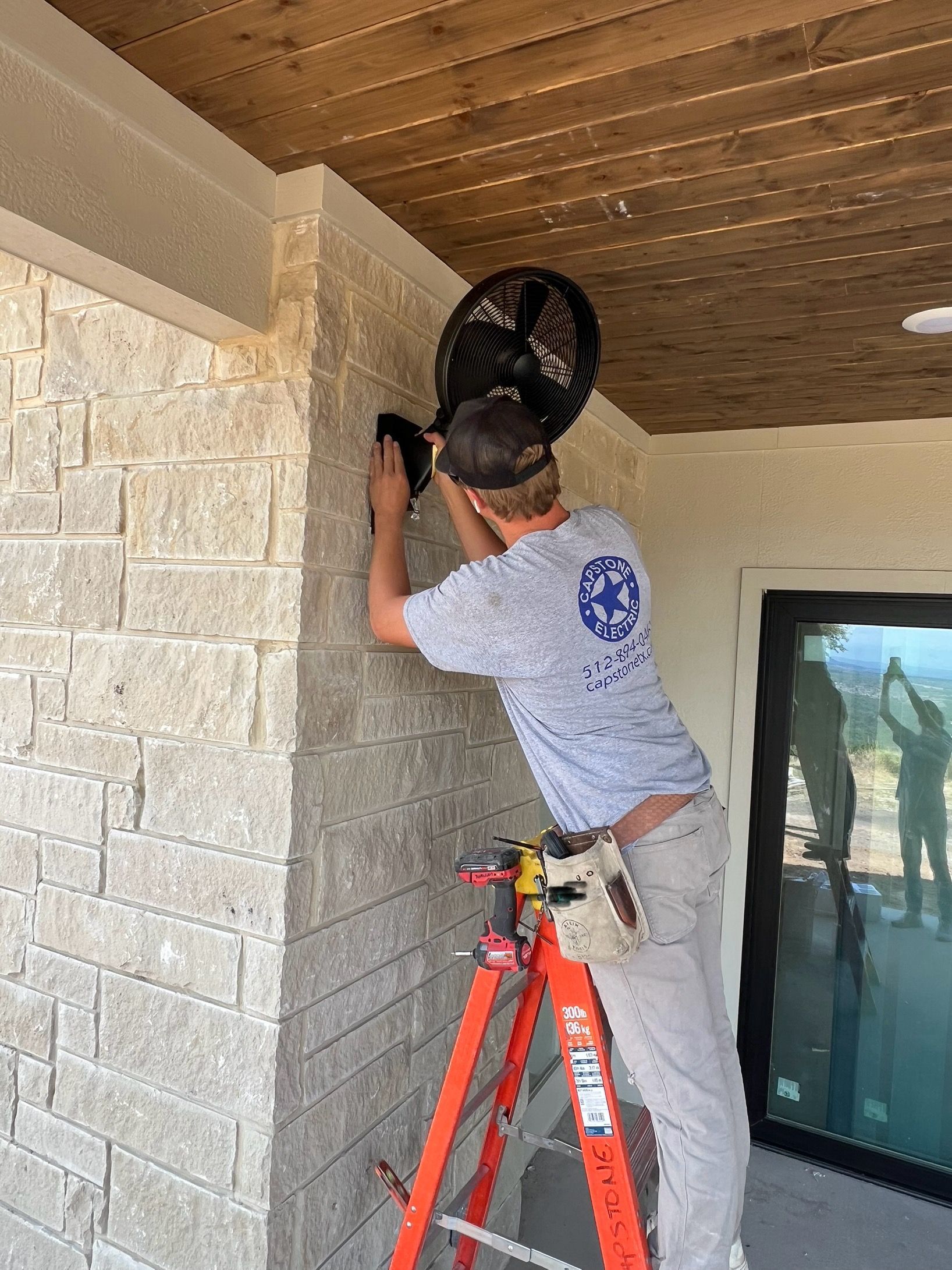 Man on ladder installing black fan on a stone wall under a wood ceiling.