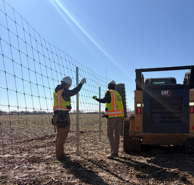 Two workers in safety vests beside a machine, installing fence in a dry field under a clear blue sky