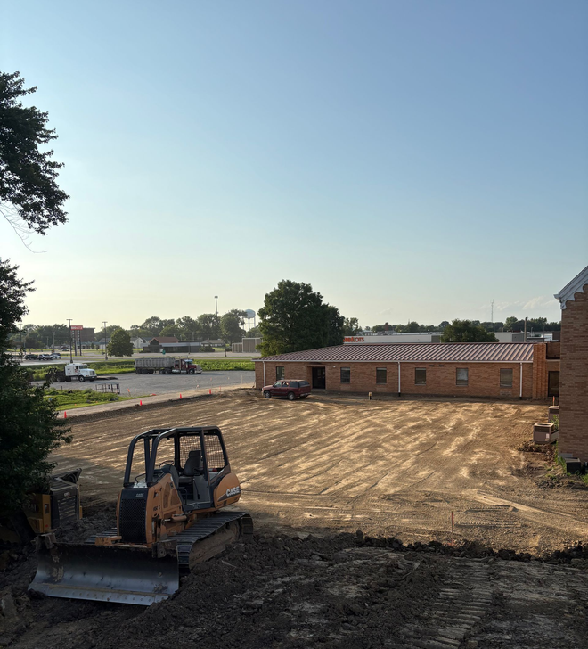 Excavator at a dirt construction site beside a brick building under a clear sky