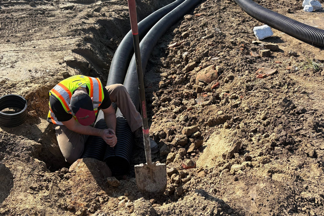 Worker in hard hat kneeling in a trench, securing black conduit on a dirt construction site.