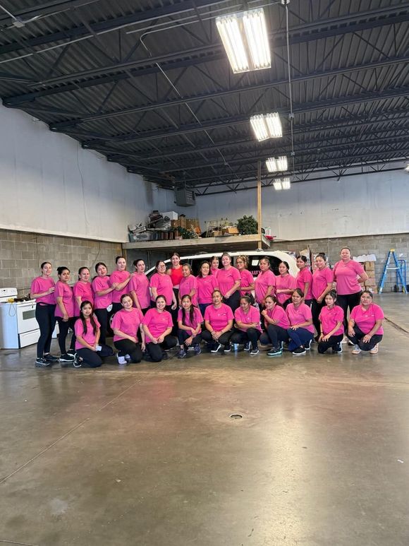Group of people in pink shirts posing in a warehouse with stacked items and concrete floors