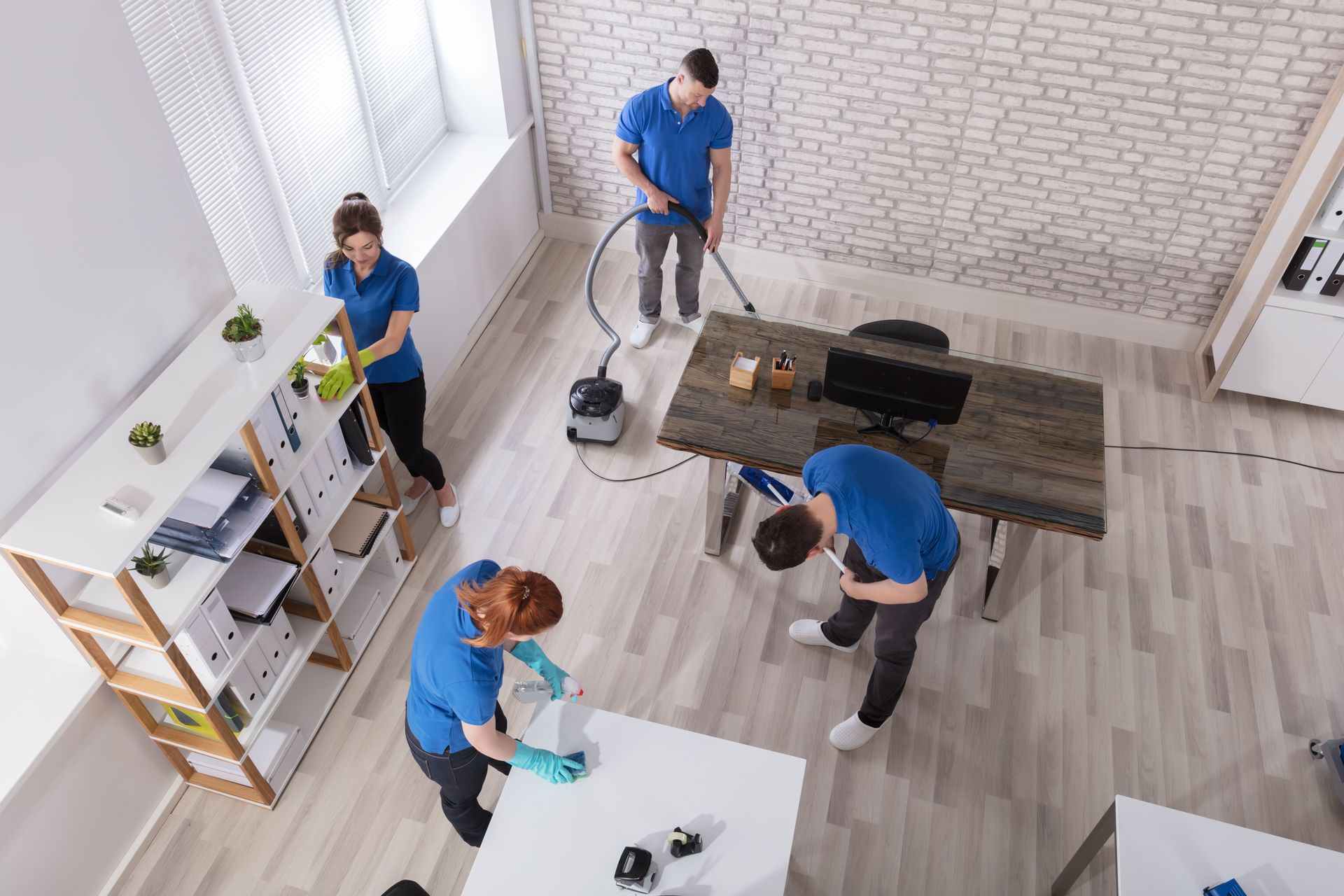 Office cleaners in blue shirts cleaning, vacuuming, dusting, and wiping surfaces.