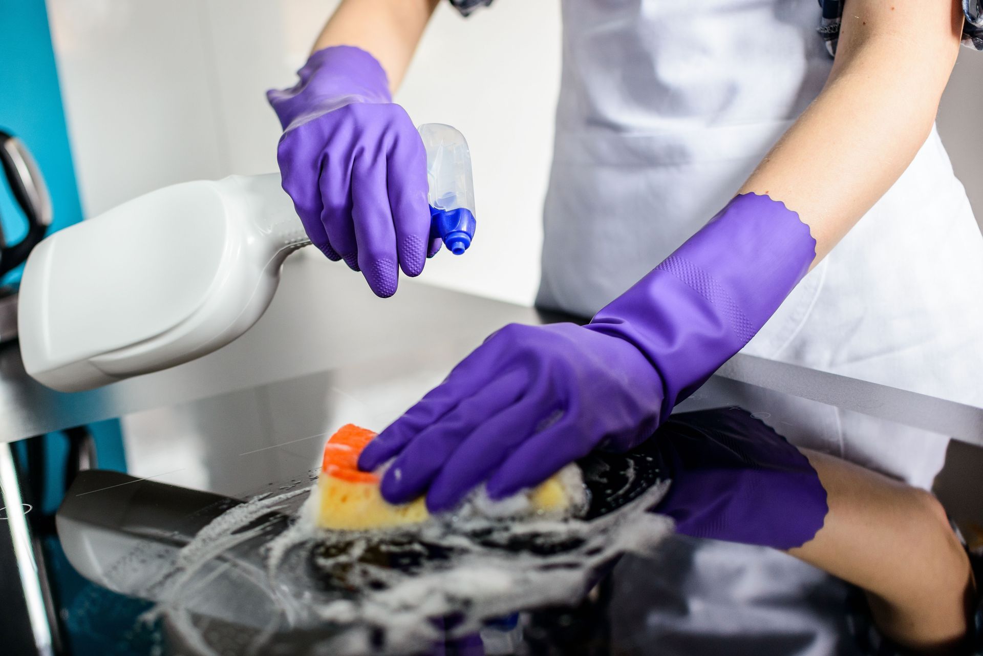 Person in purple gloves cleaning a dark sink with a sponge and spray bottle.