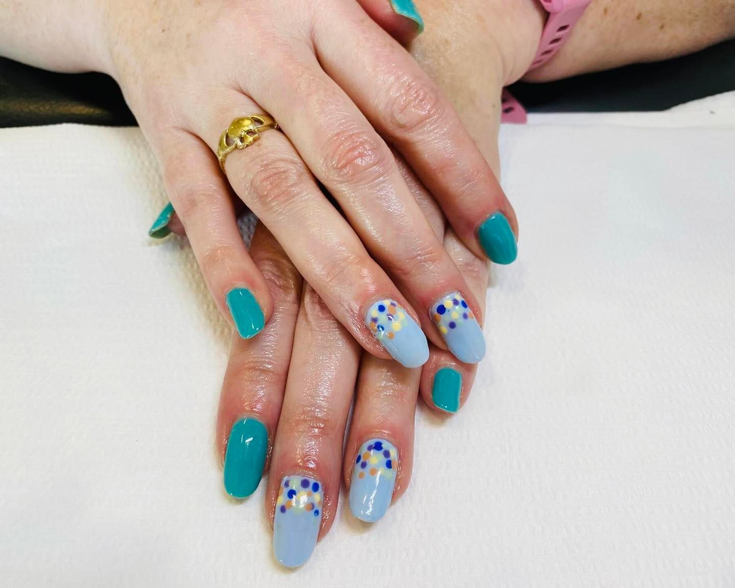 Hands with teal and light blue, decorated nails. One hand has a gold ring.