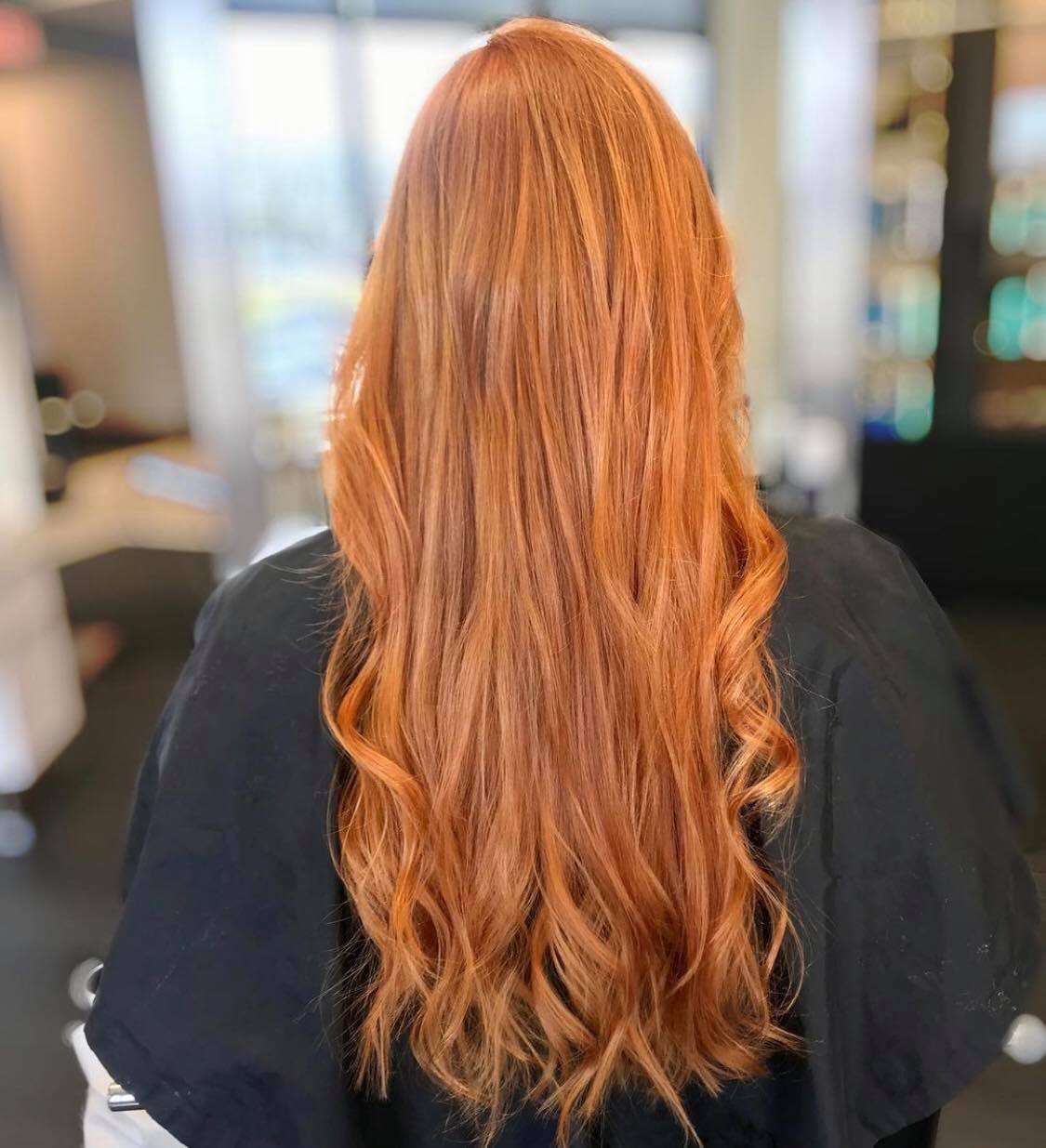 Woman with long, wavy, vibrant orange hair, viewed from the back, in a salon.