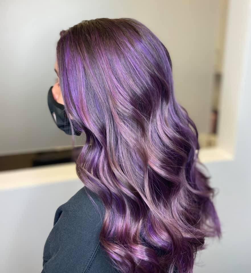 Woman with long, wavy, purple hair. Wearing a black face mask and black top.