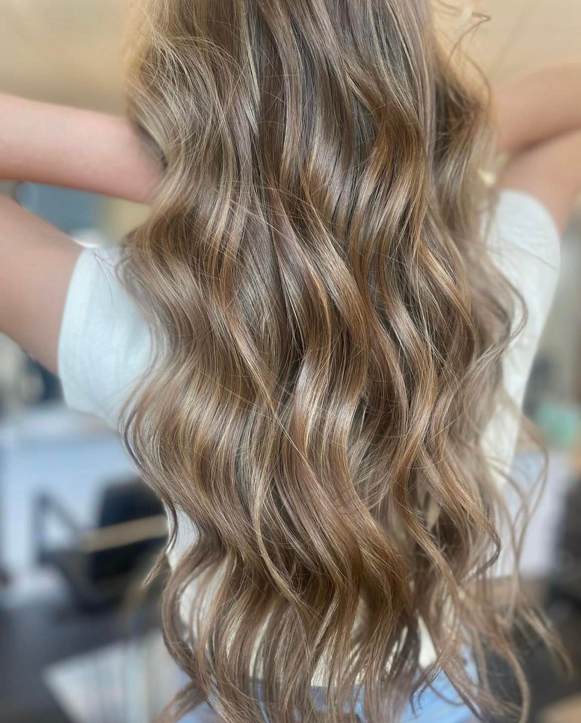Woman with long, wavy, highlighted brown hair, arms raised, wearing a white shirt.
