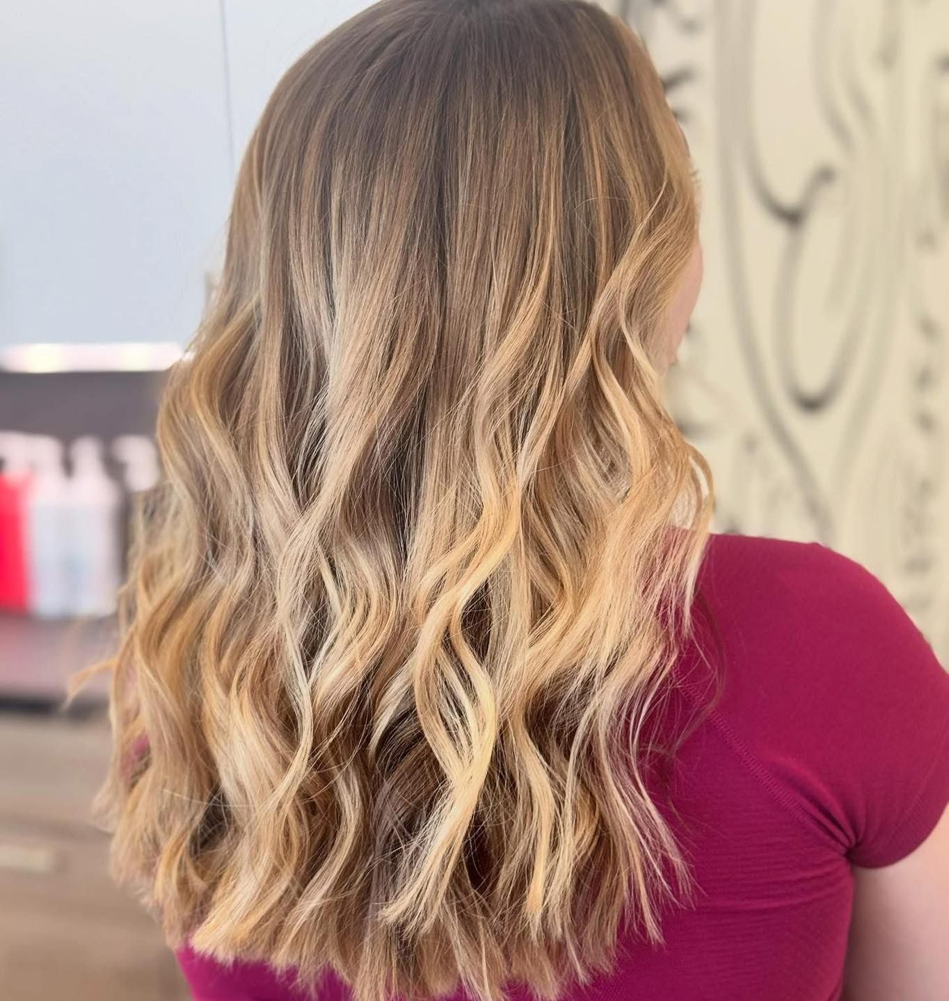 Woman with long, wavy, blonde-ombre hair styled at a salon, wearing a burgundy top.