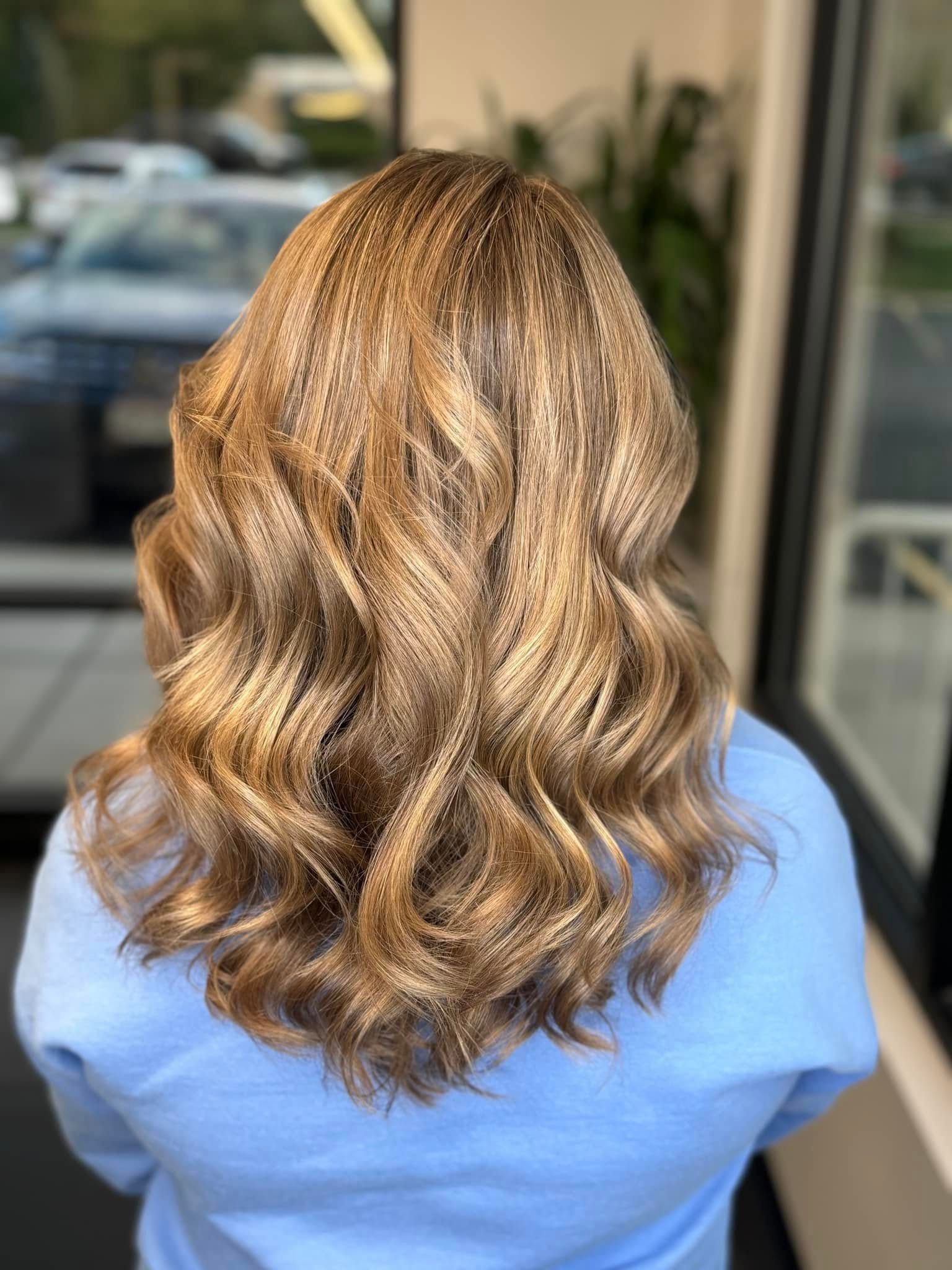 Woman with wavy, crimped light brown hair, wearing a blue sweater, in a salon.