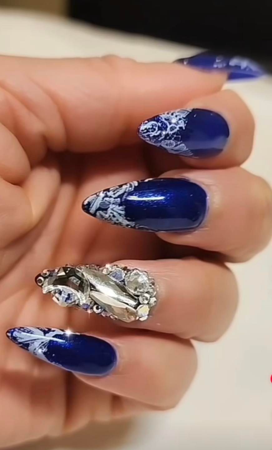Hand with long, pointed navy blue nails, some with white lace design, one with large silver rhinestones.
