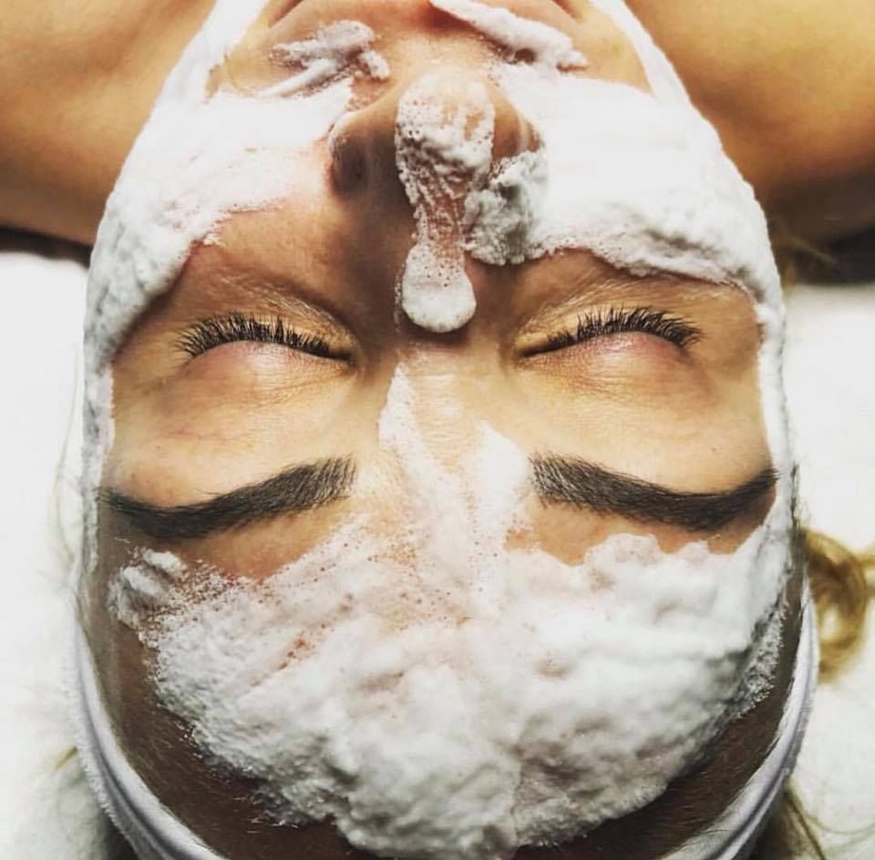 Woman with closed eyes, covered in a facial foam mask, lying on a white surface.