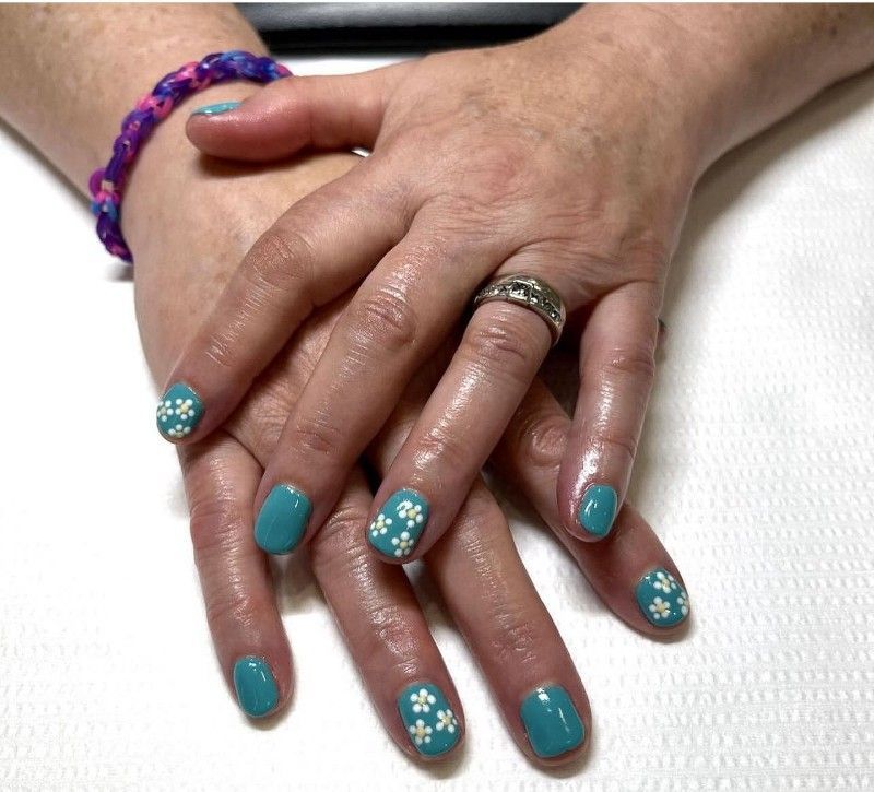 Hands with turquoise nails, each adorned with white floral designs. A silver ring and a purple bracelet are also visible.