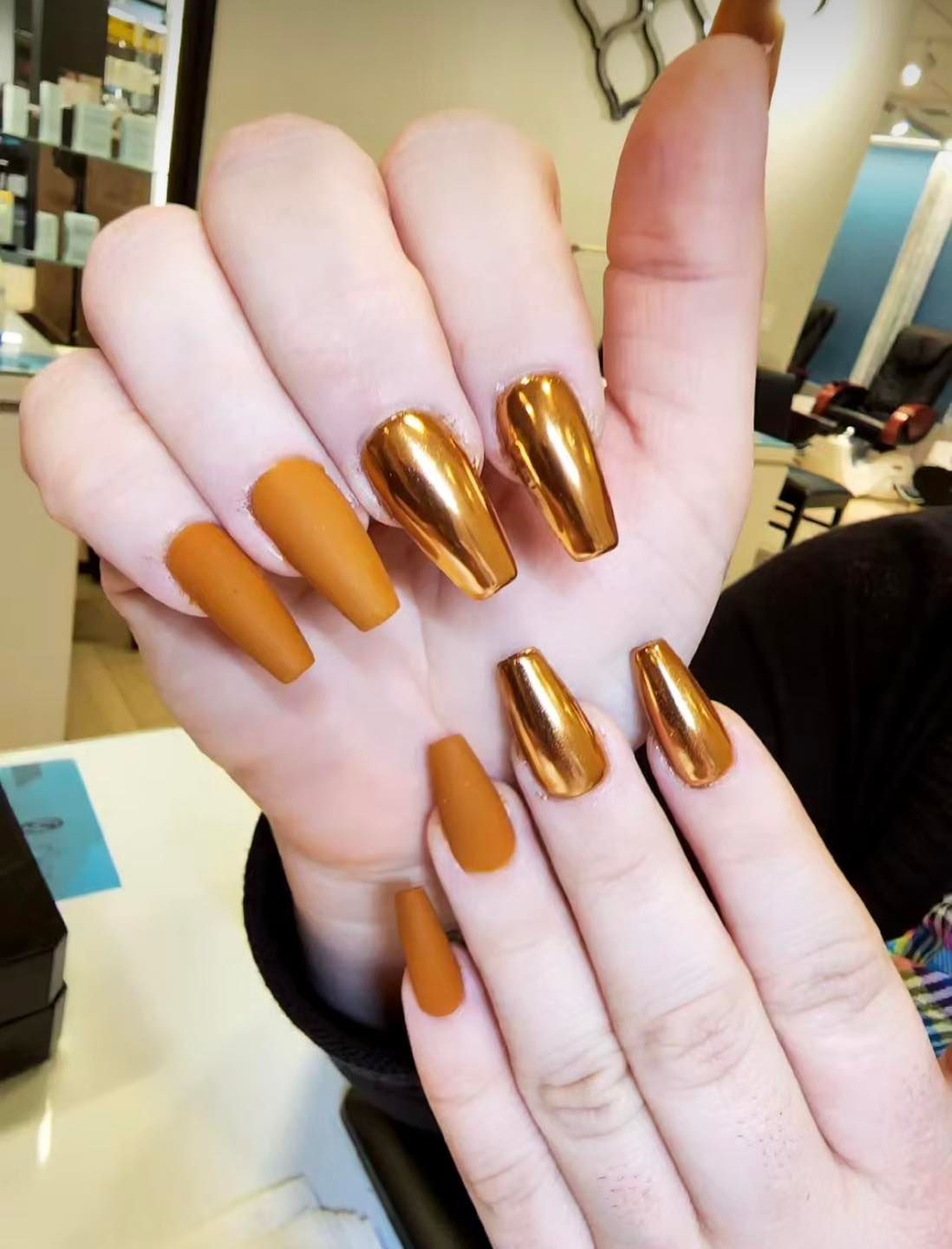 Hands displaying coffin-shaped nails: matte orange and chrome gold, in a beauty salon setting.