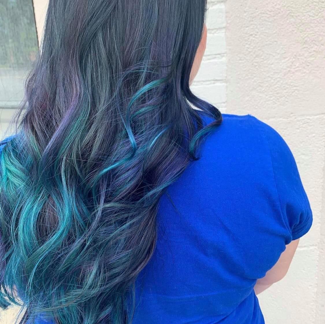 Woman with long dark hair, teal and purple highlights, wearing a blue shirt, standing outdoors.