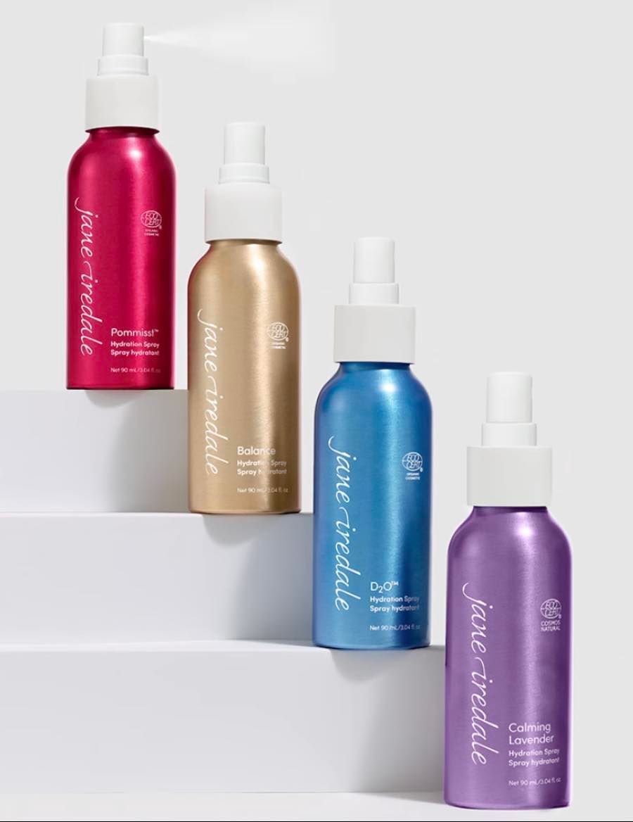 Four colorful jane iredale facial sprays in metallic bottles, arranged on steps, against a white background.