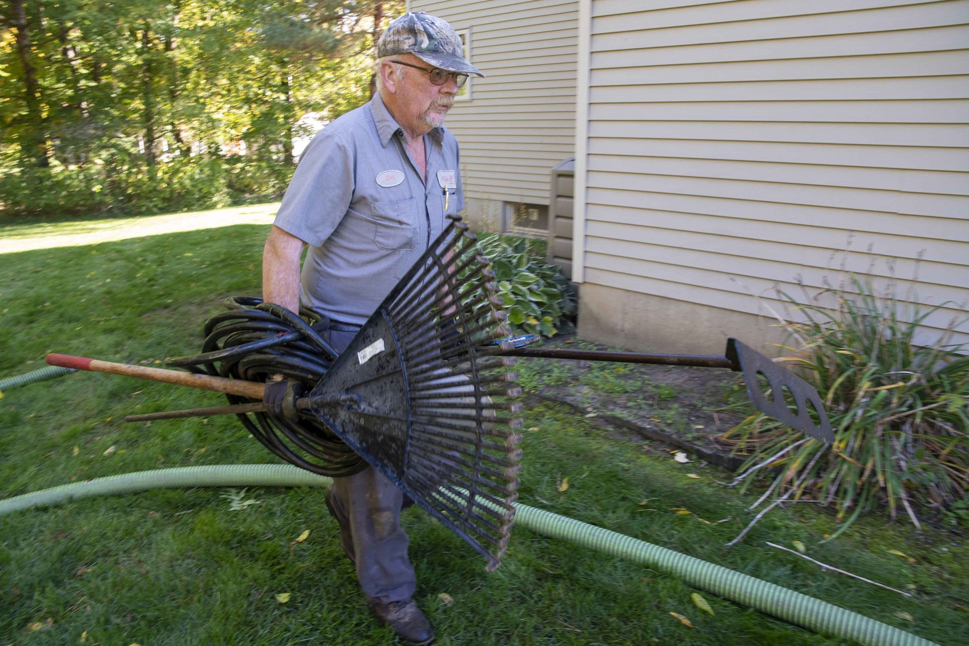 Man carrying rake, hose, and other tools near a house on a sunny day.