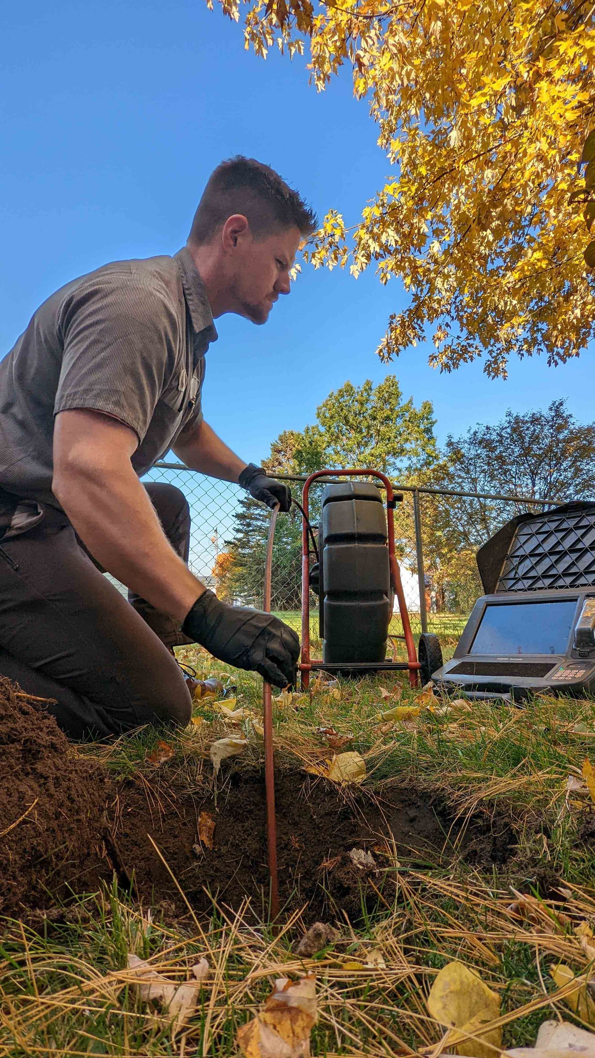 Man kneels to install a grounding rod outdoors. A rolling machine and computer sit nearby.